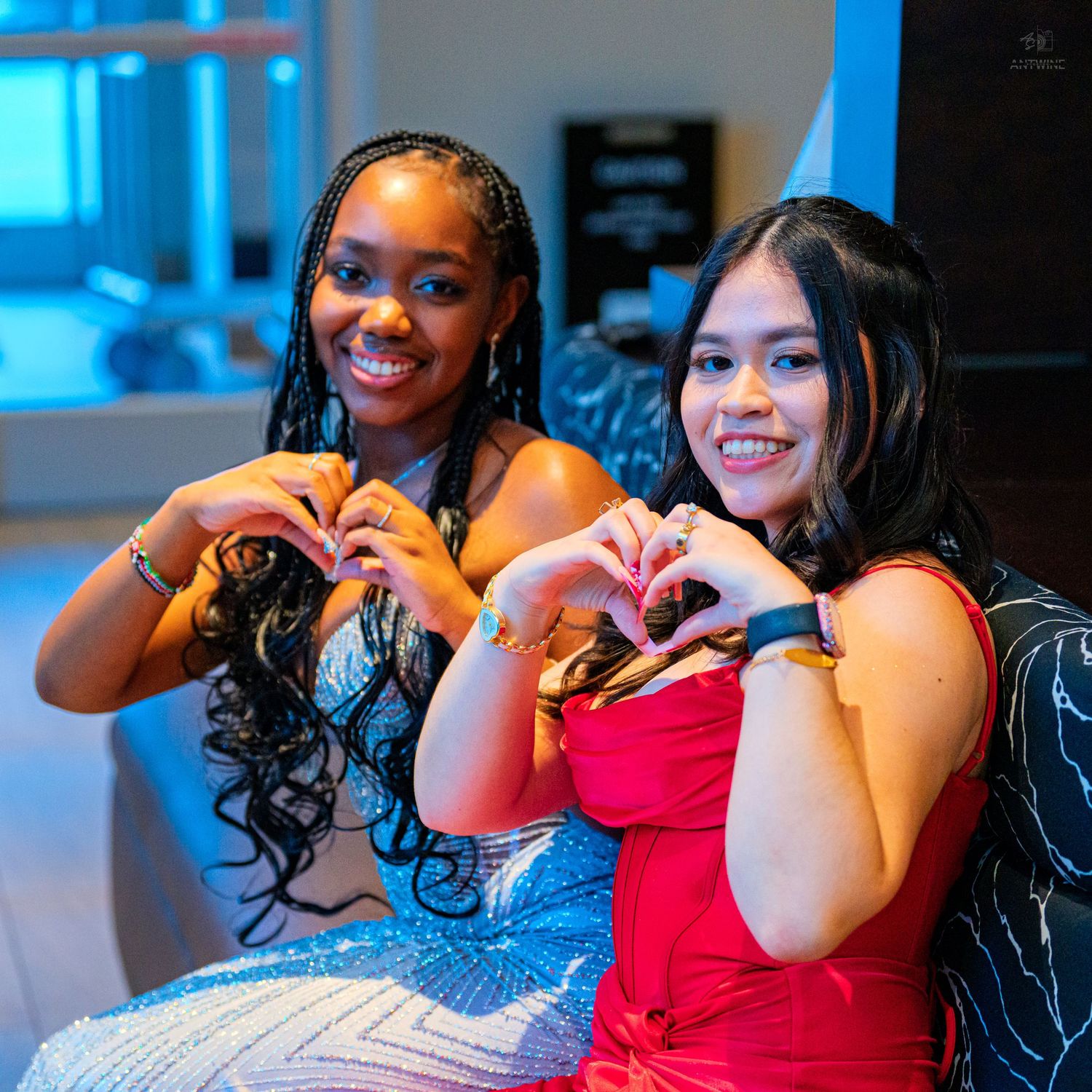 Two young women in formal dresses making heart shapes with their hands while smiling at a party.