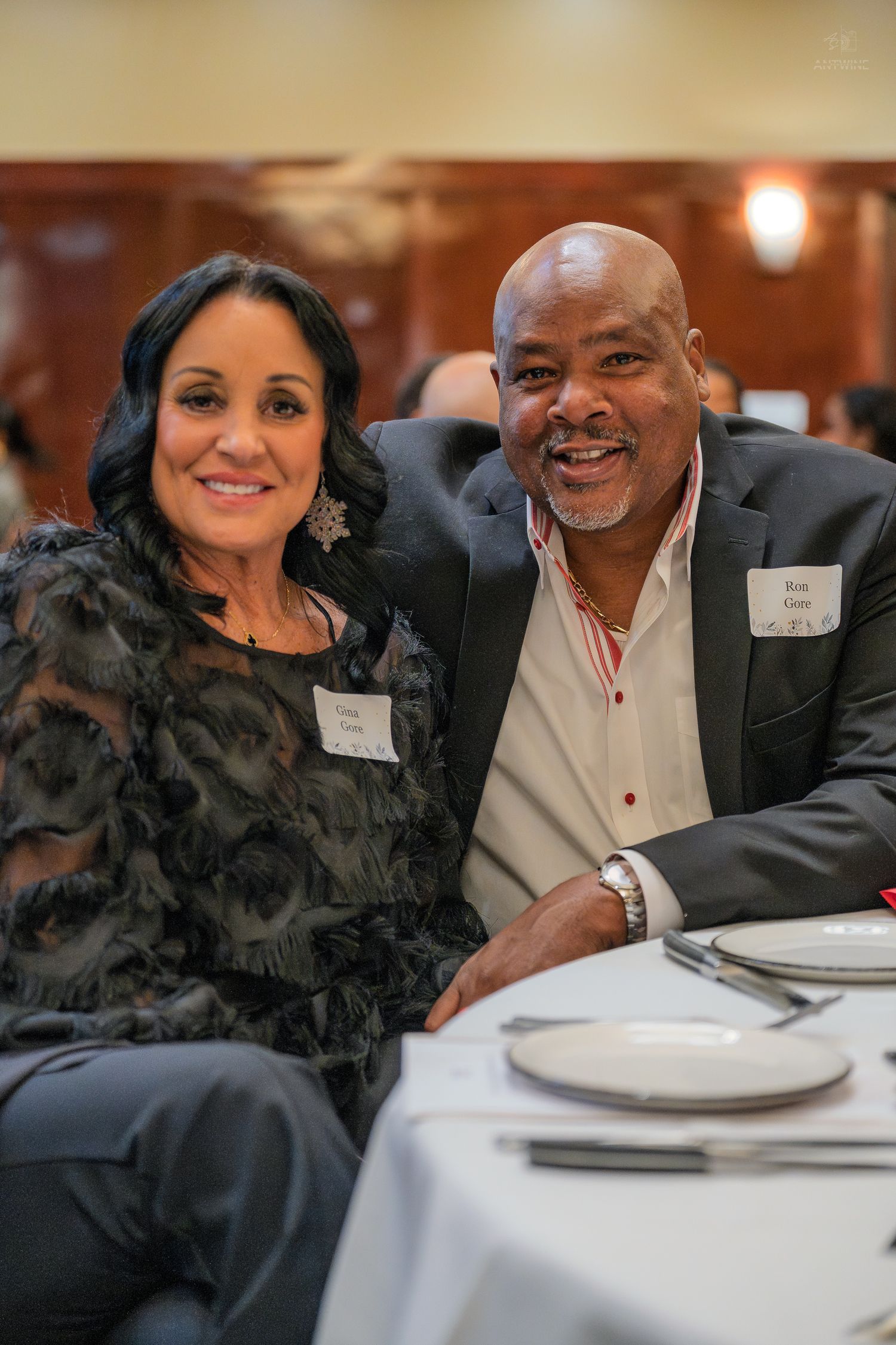 Smiling couple poses together at a formal dinner event, wearing name tags and seated at a white tablecloth table.