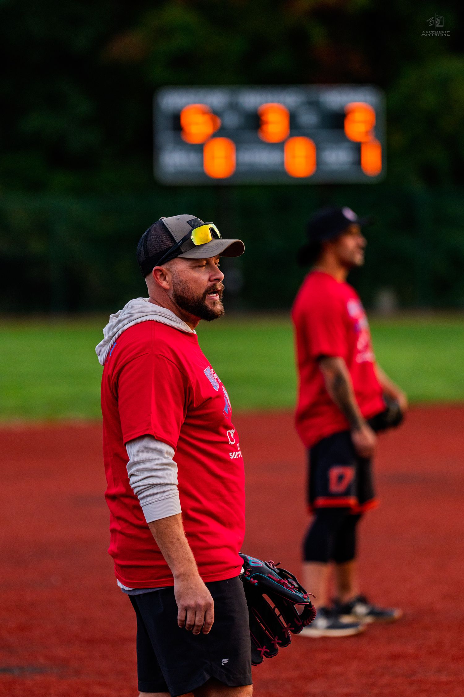 Two men in red shirts on softball field, one holding glove with scoreboard lit up behind them.
