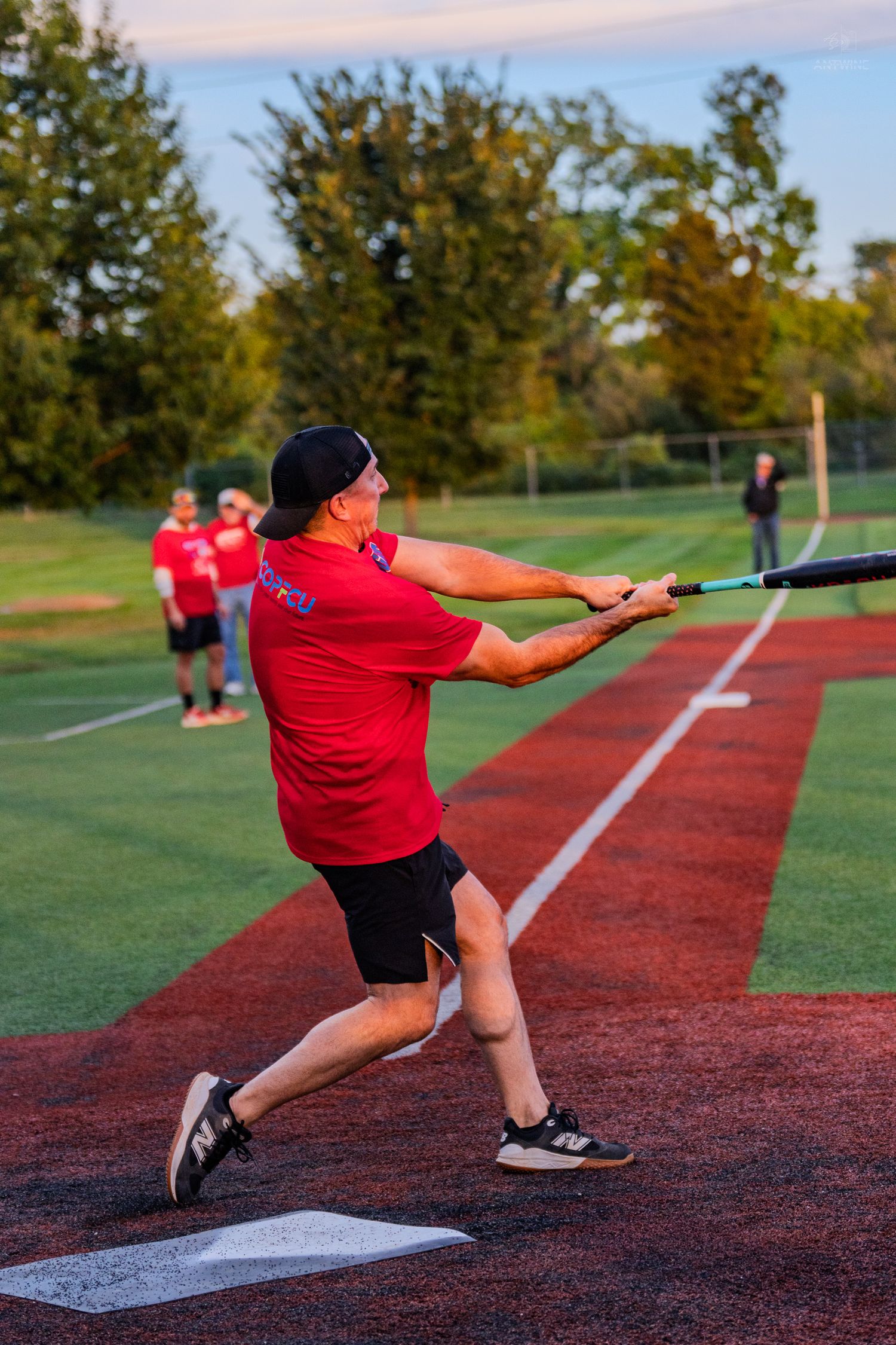 A batter in a red shirt swings a baseball bat during a softball game on a red turf field at sunset.