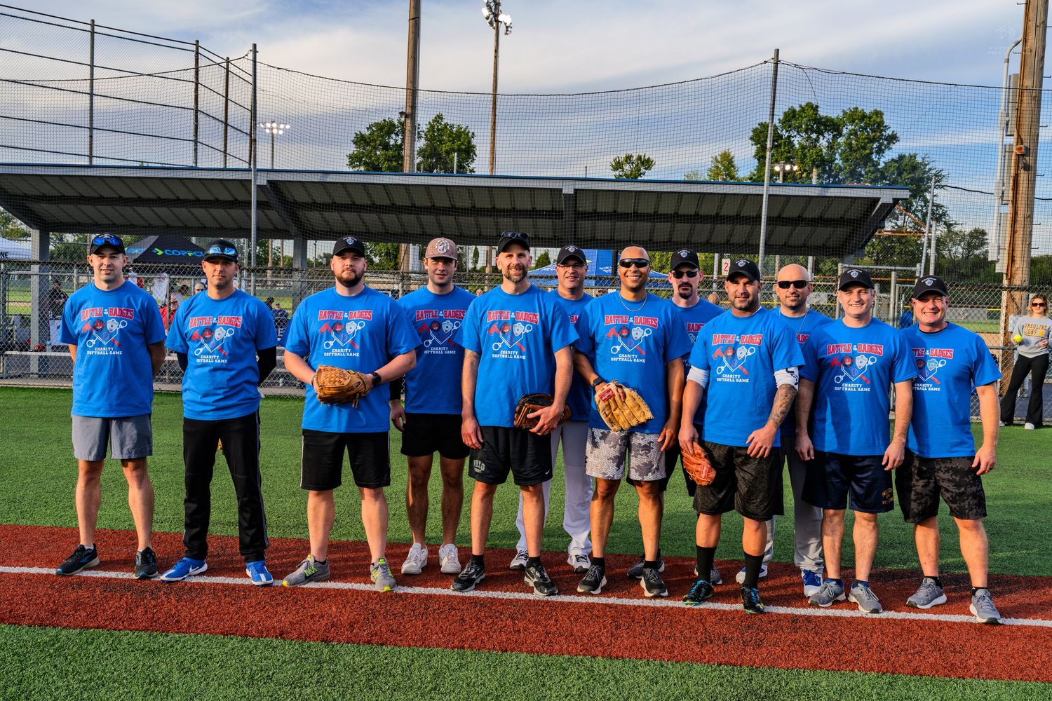A softball team in blue matching shirts poses together on a baseball diamond during a clear evening.