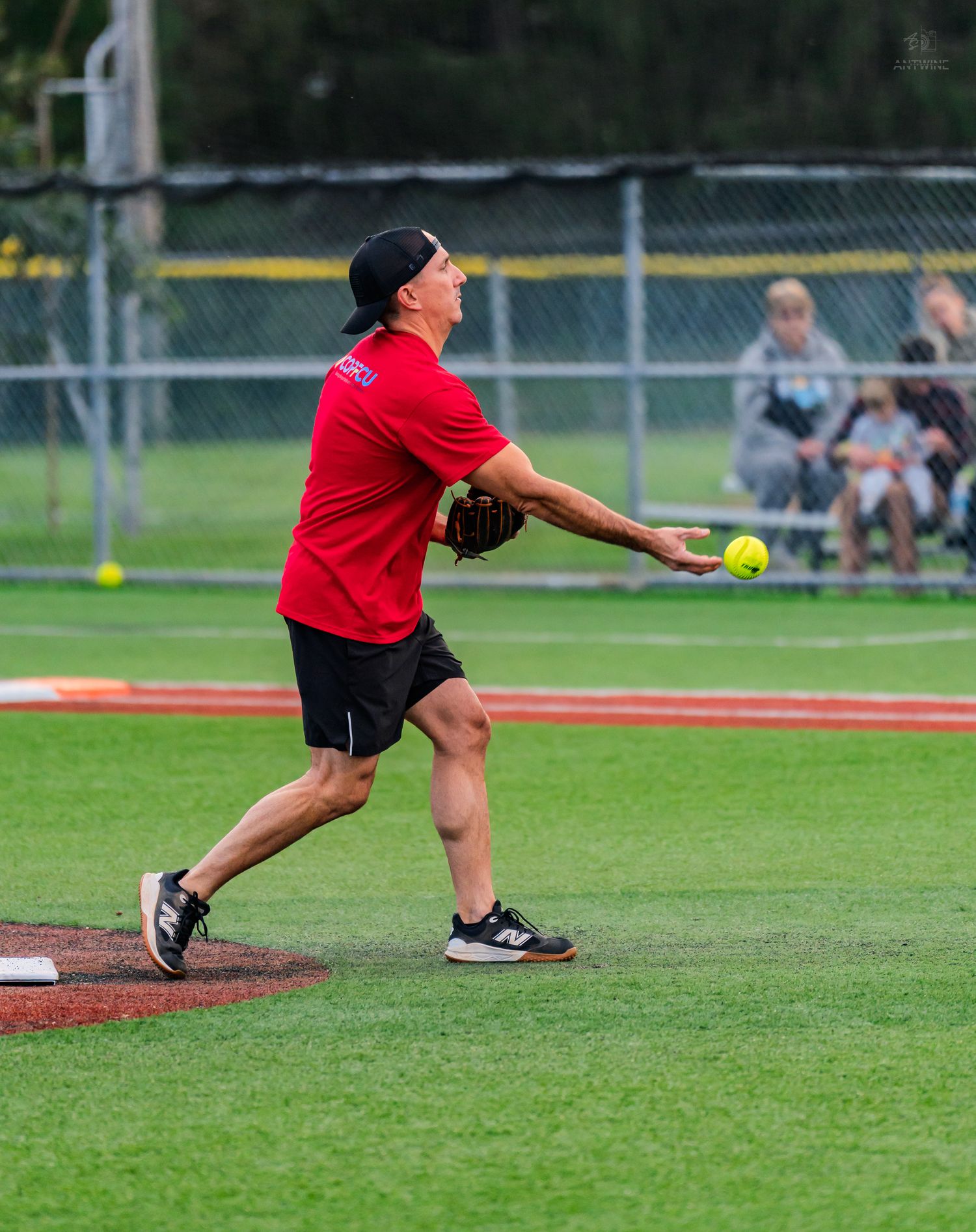 An athlete in a red shirt fields a yellow softball on a pitchers mound during an outdoor game.