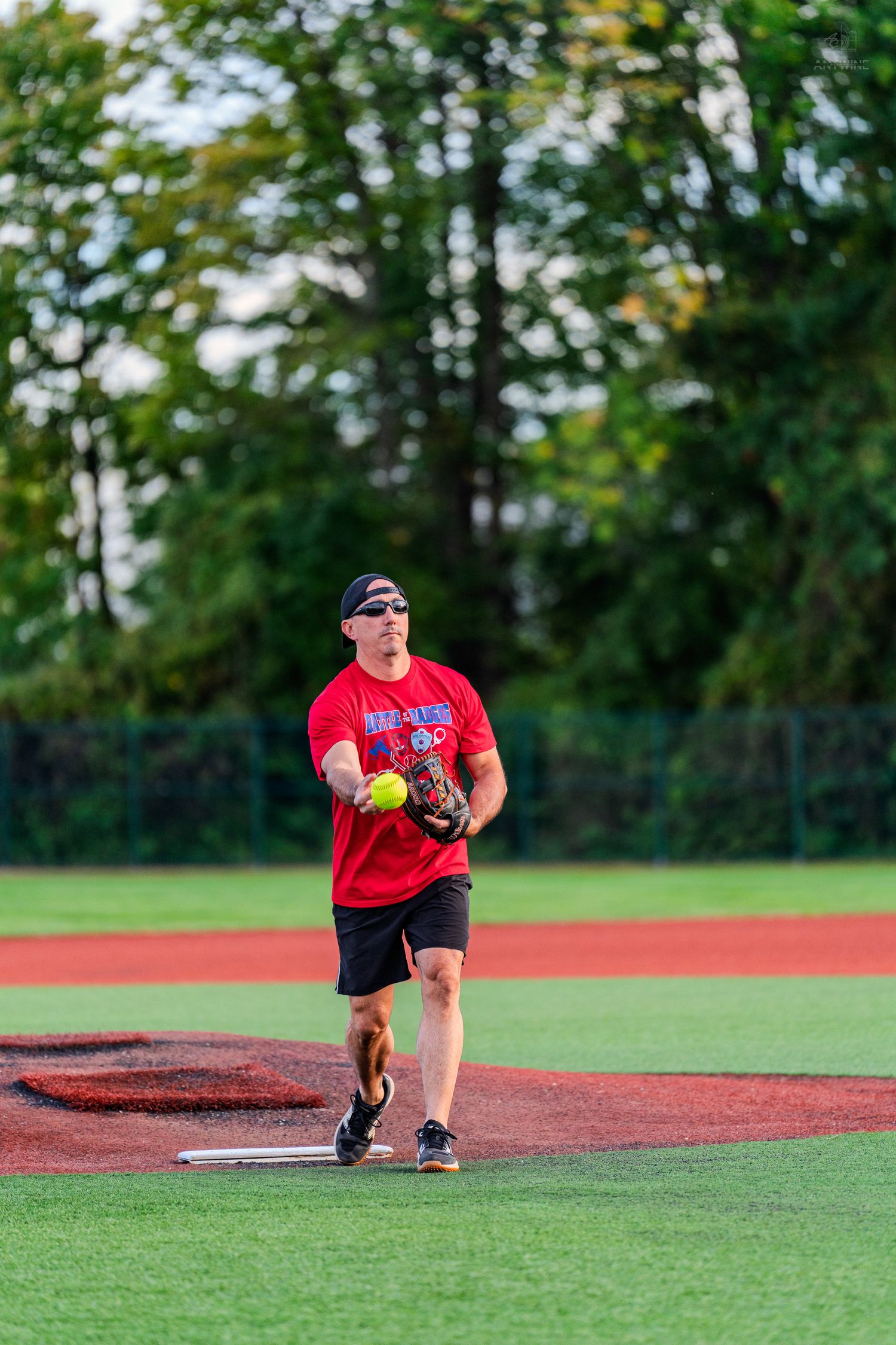 Man in red shirt pitching a yellow softball on a green field during an outdoor recreational softball game.