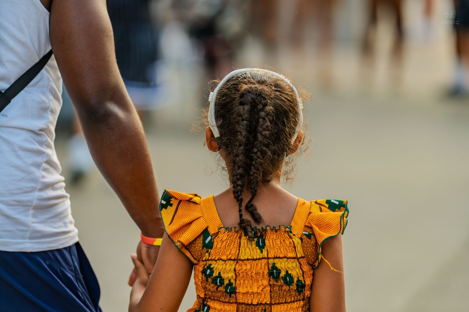 Young girl in colorful African print dress holds adult's hand while walking outdoors.
