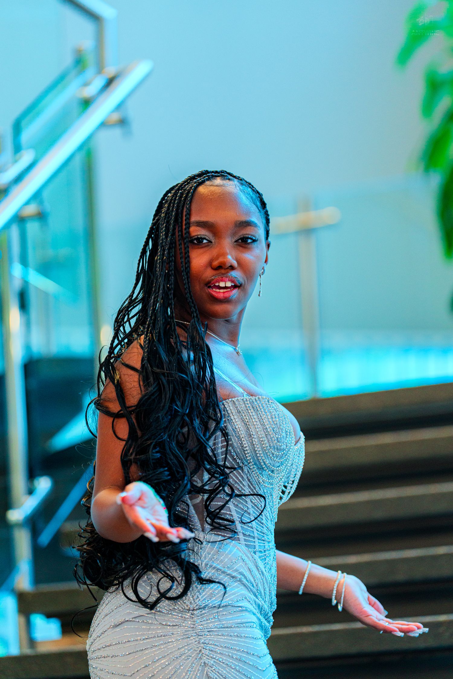 Young Black woman with long box braids in a silver dress poses elegantly near modern glass staircase.