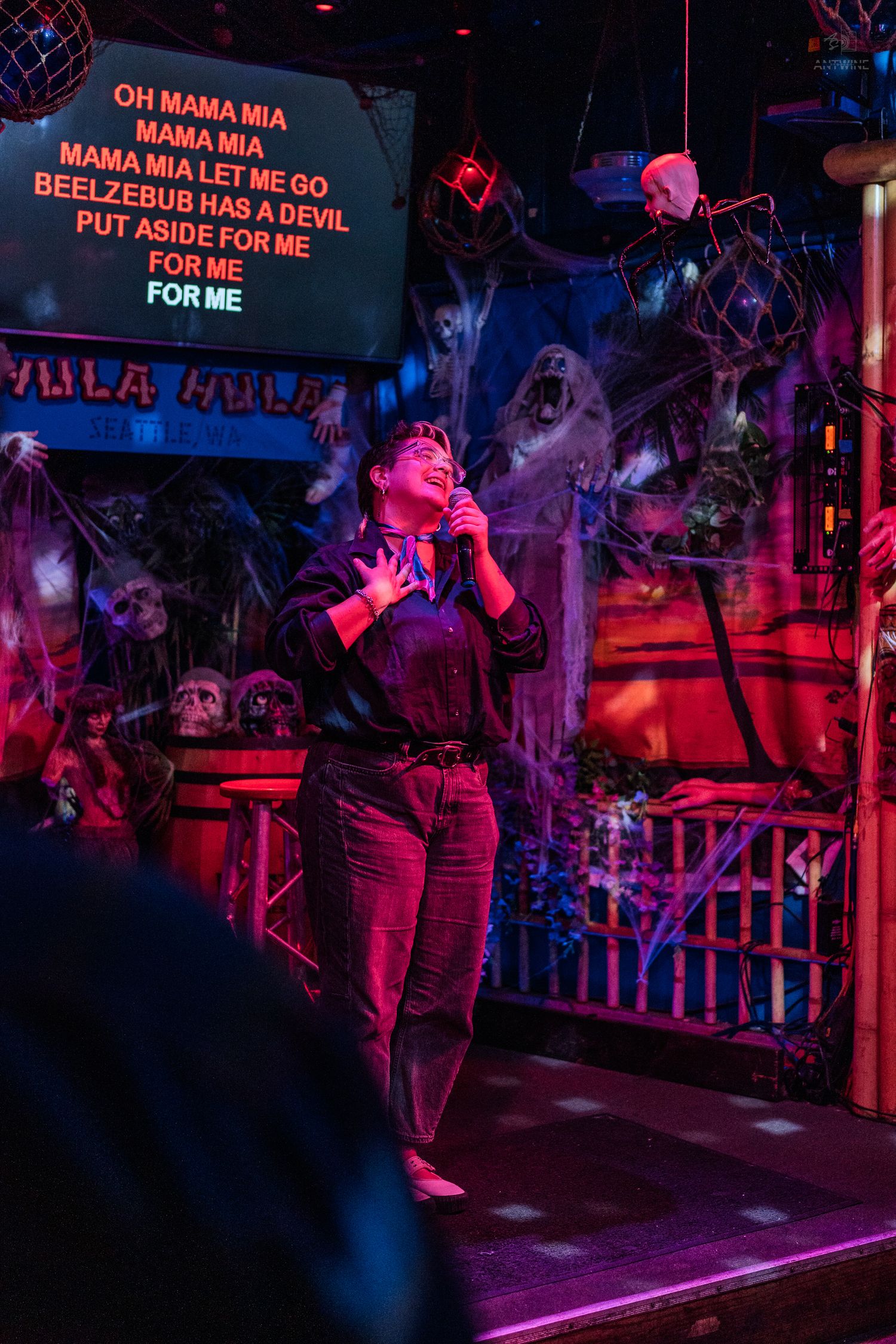 Person singing karaoke to Bohemian Rhapsody in a Halloween-decorated bar with red lighting.