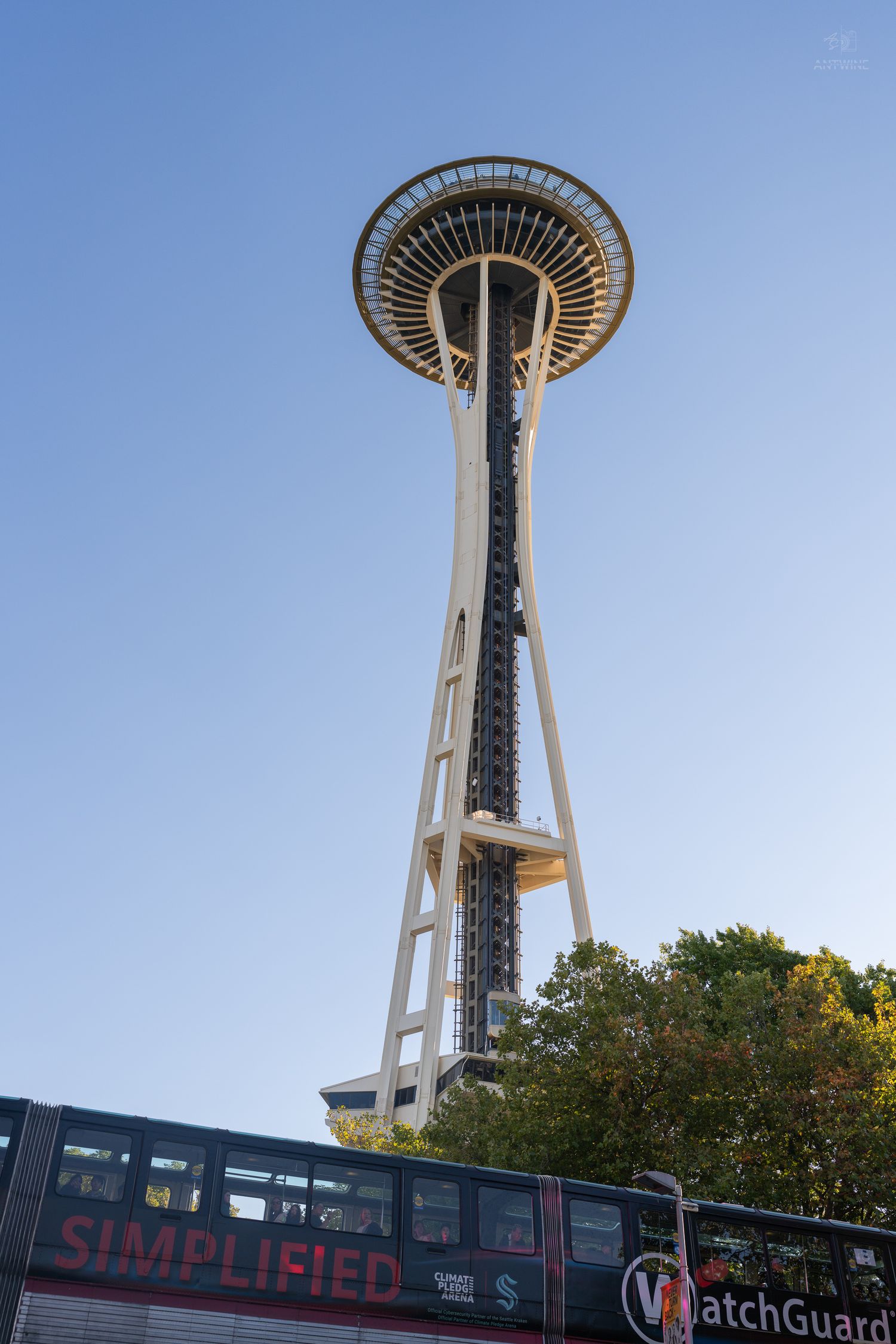 Seattle Space Needle viewed from below against blue sky with a WatchGuard bus and trees visible below.