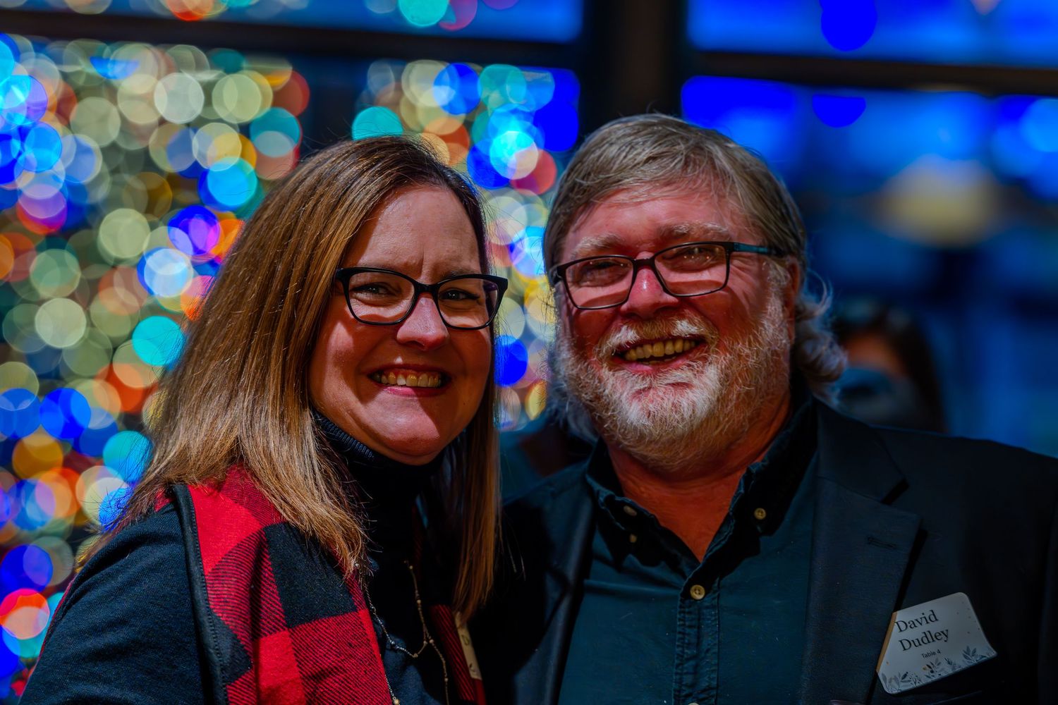 A smiling couple poses together in front of colorful bokeh Christmas lights at a holiday party.