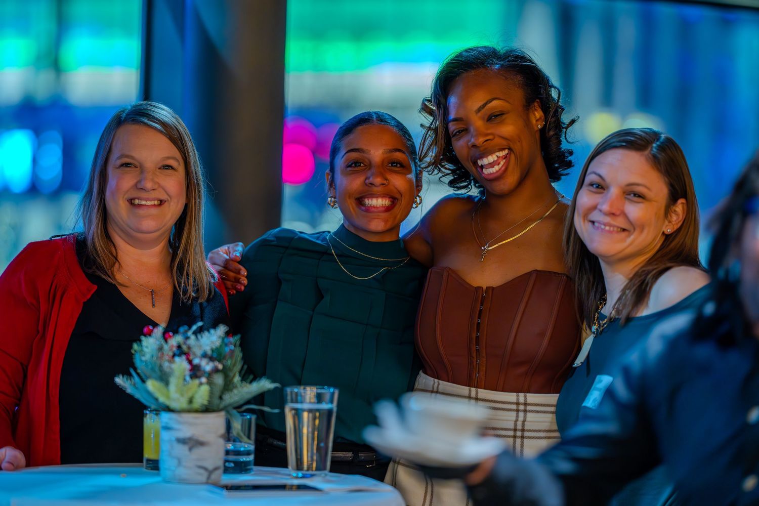 Four smiling women pose together at a dinner event, with colorful blue and green lights in the background.