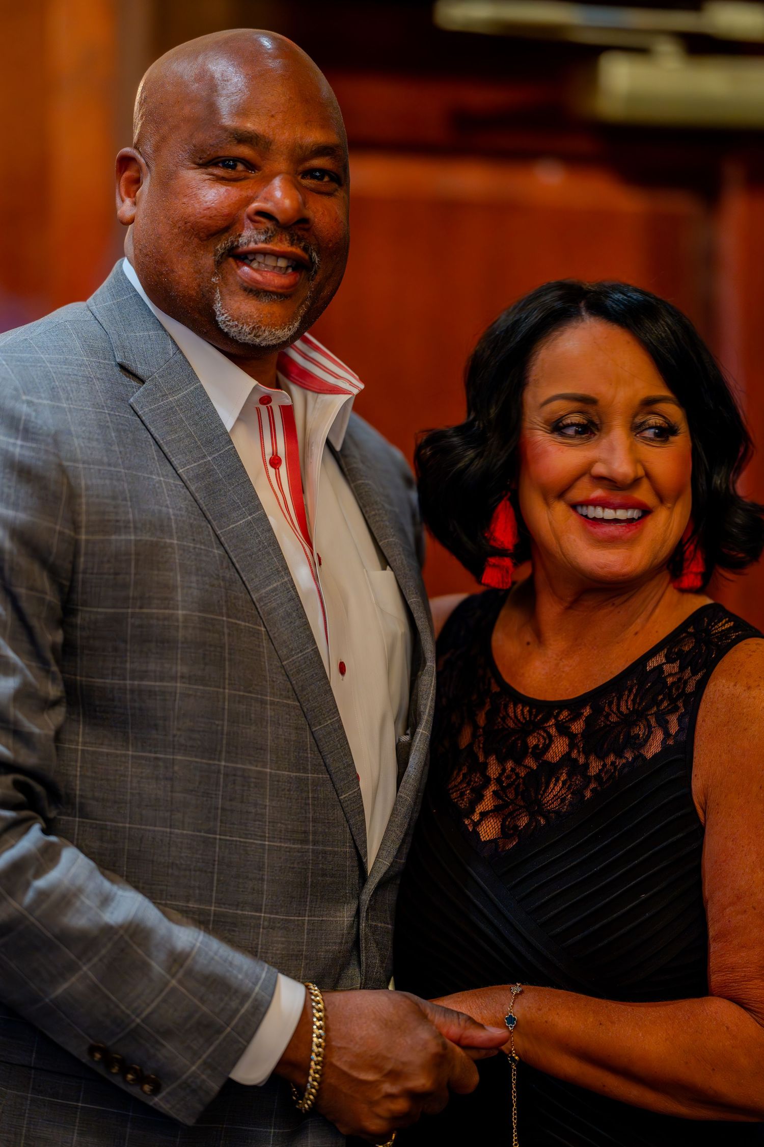 Smiling couple dressed elegantly, man in gray suit and woman in black lace dress, posing together at a formal event.