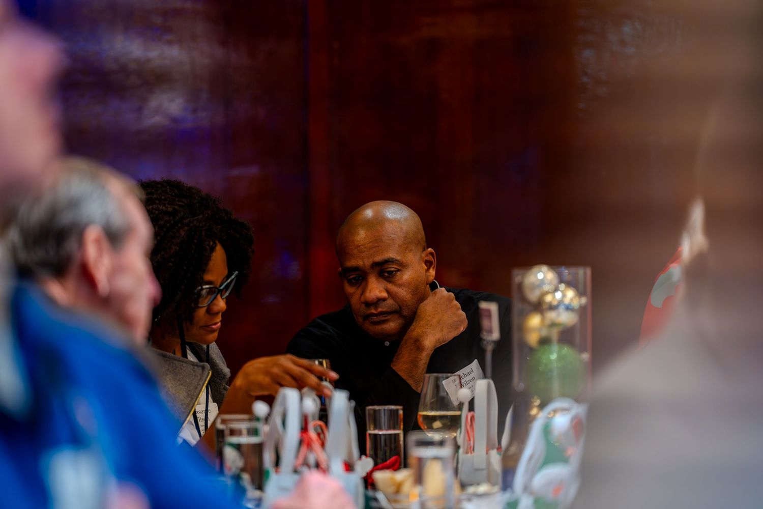 Two people engaged in conversation at a dinner table with glasses and decorations in a dimly lit restaurant.