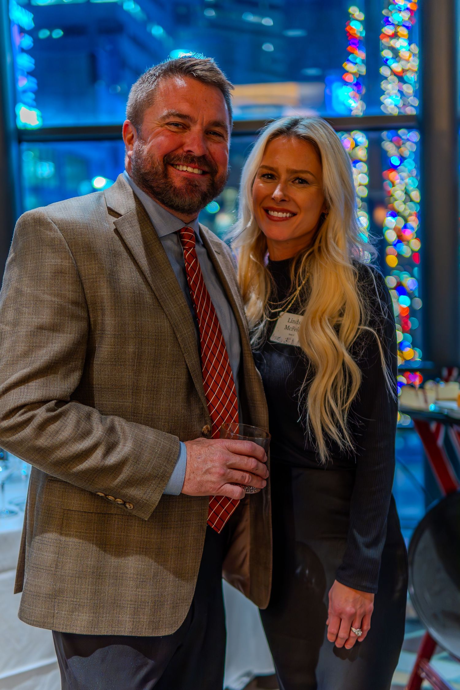 A smiling couple poses together at a festive holiday party with colorful bokeh lights in the background.