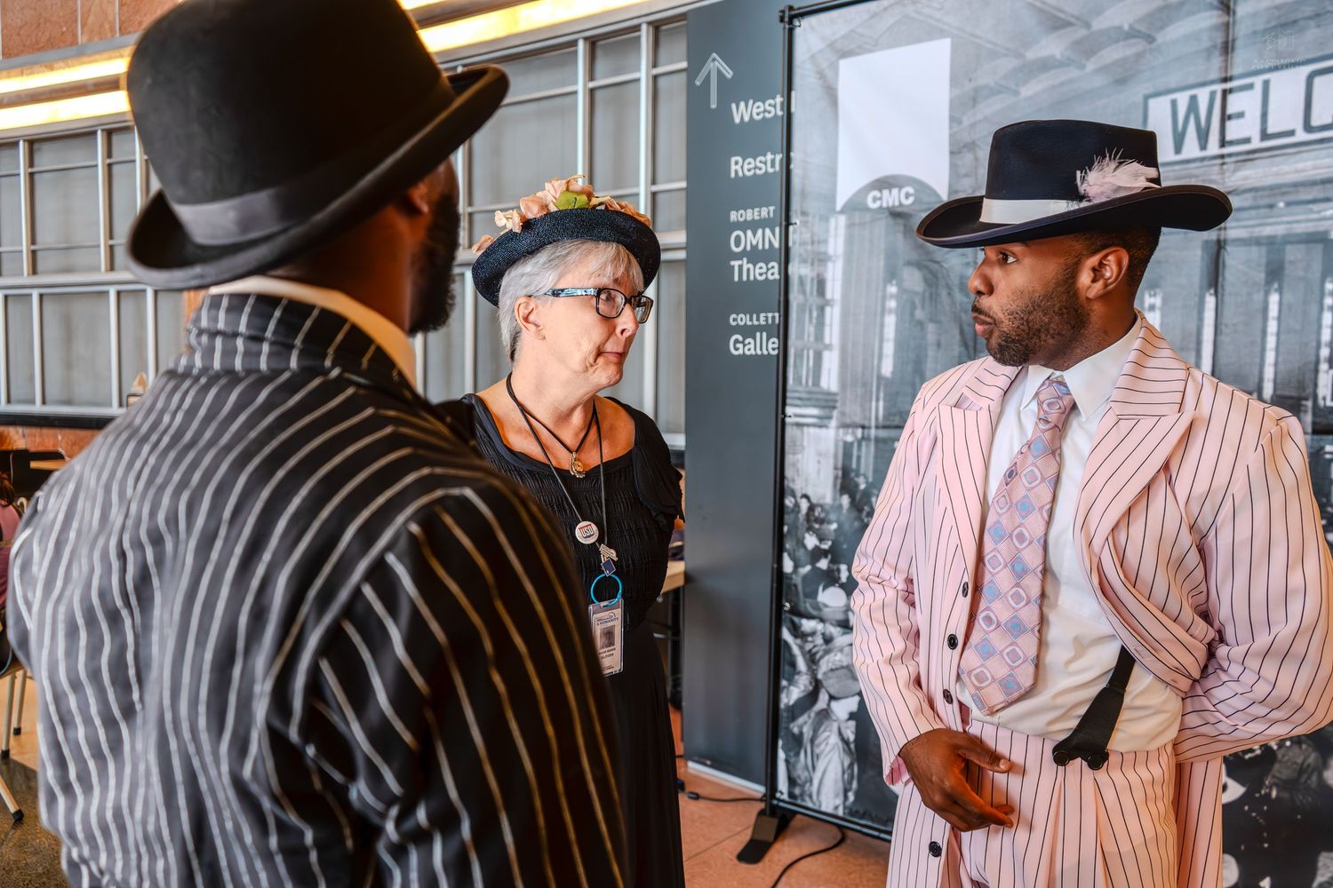 Three people in vintage zoot suits and wide-brim hats converse at a historical exhibit display.