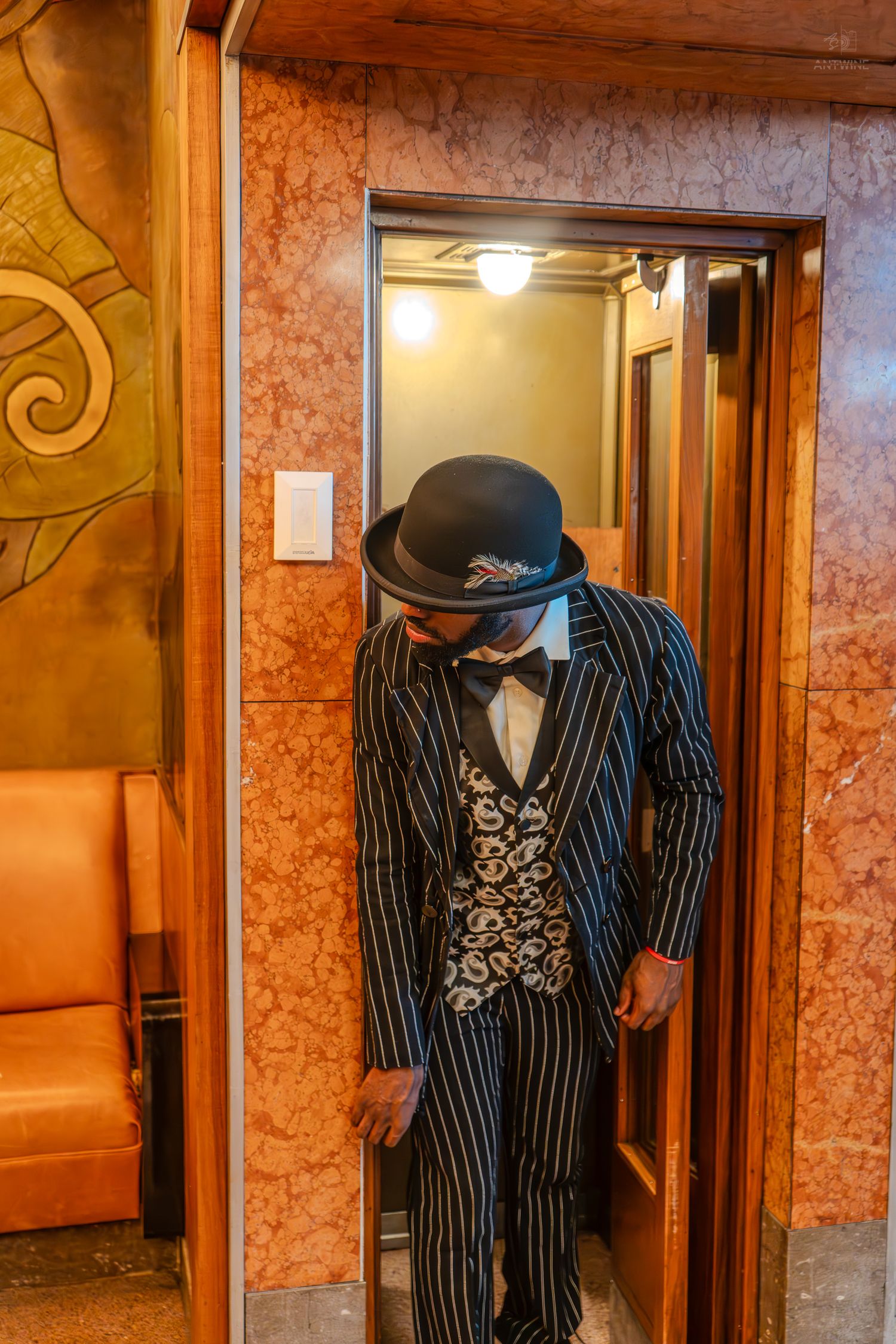 Man in pinstripe suit, bowler hat, and bow tie standing in an ornate elevator doorway with marble walls.