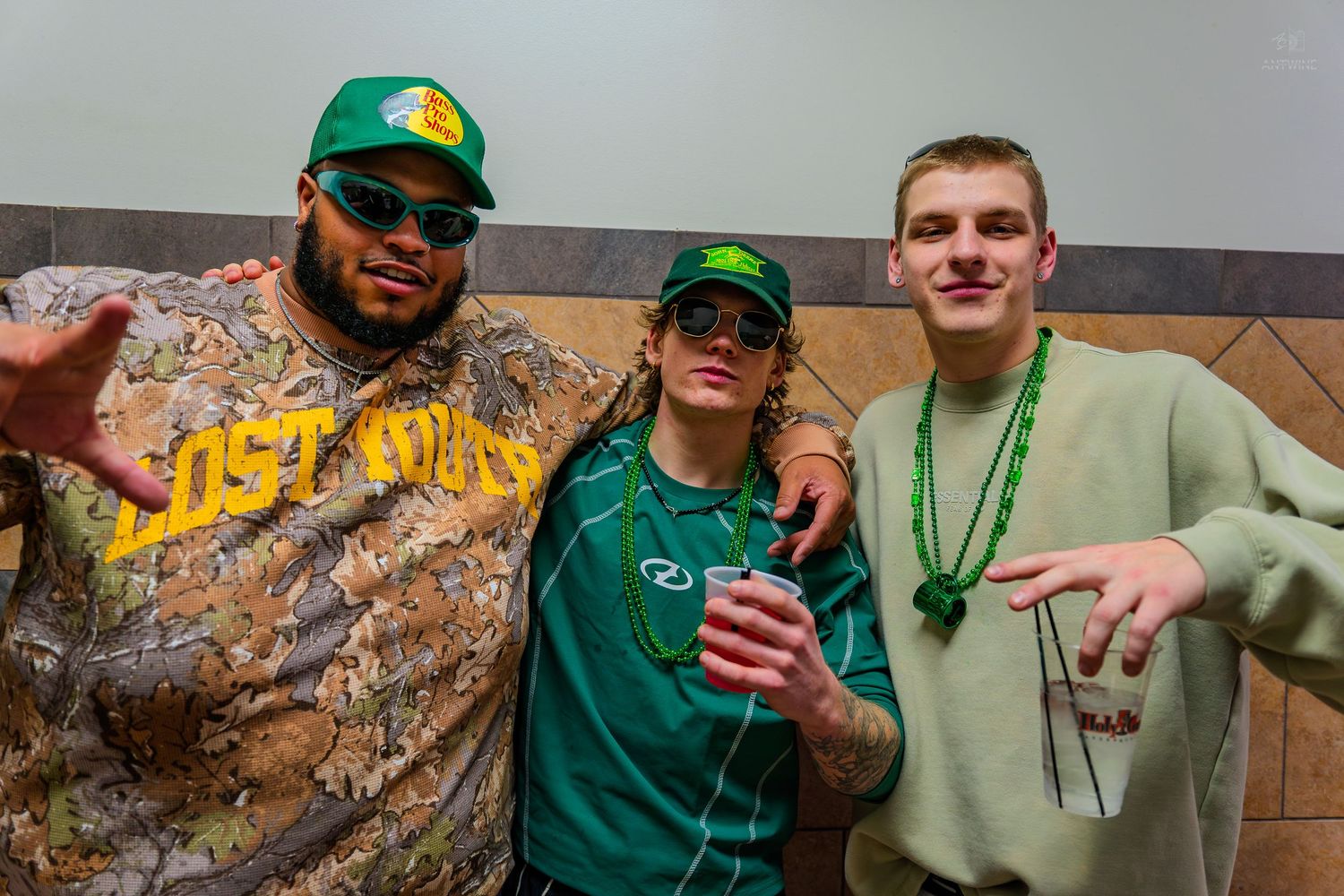Three friends pose together wearing green St. Patrick's Day outfits, hats, and beaded necklaces at a festive celebration.
