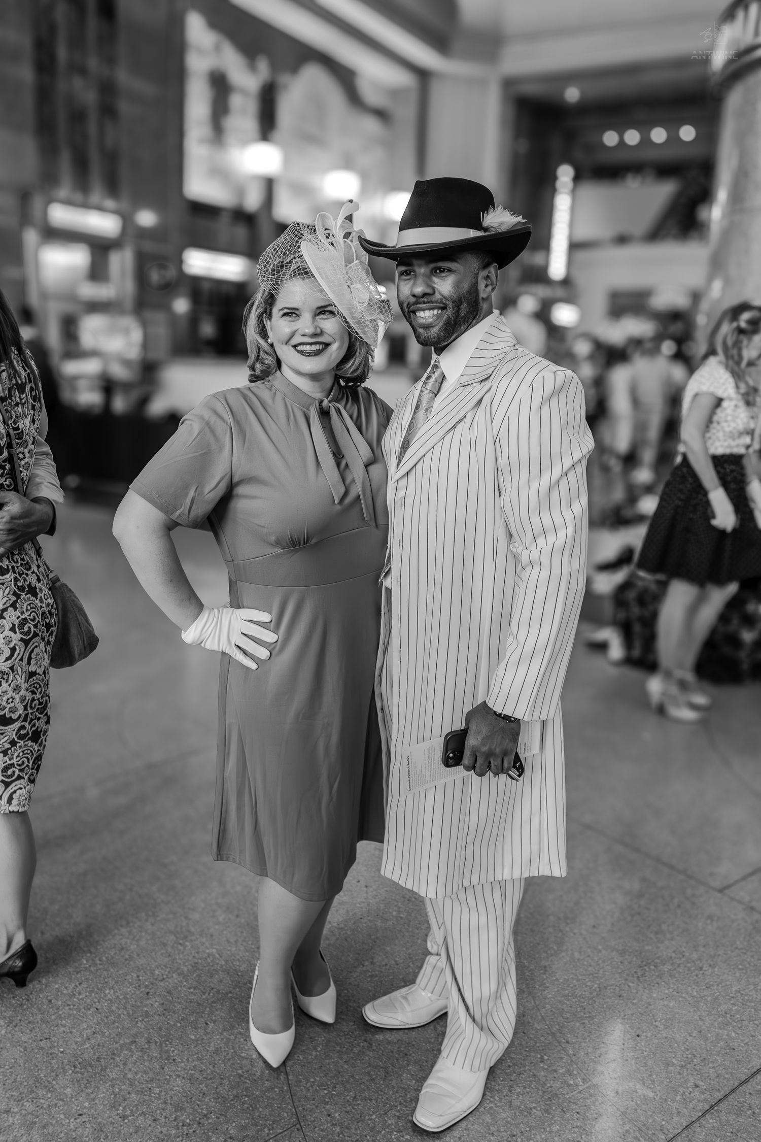 Black and white photo of a couple in 1940s vintage attire, woman in dress with fascinator, man in pinstripe zoot suit.