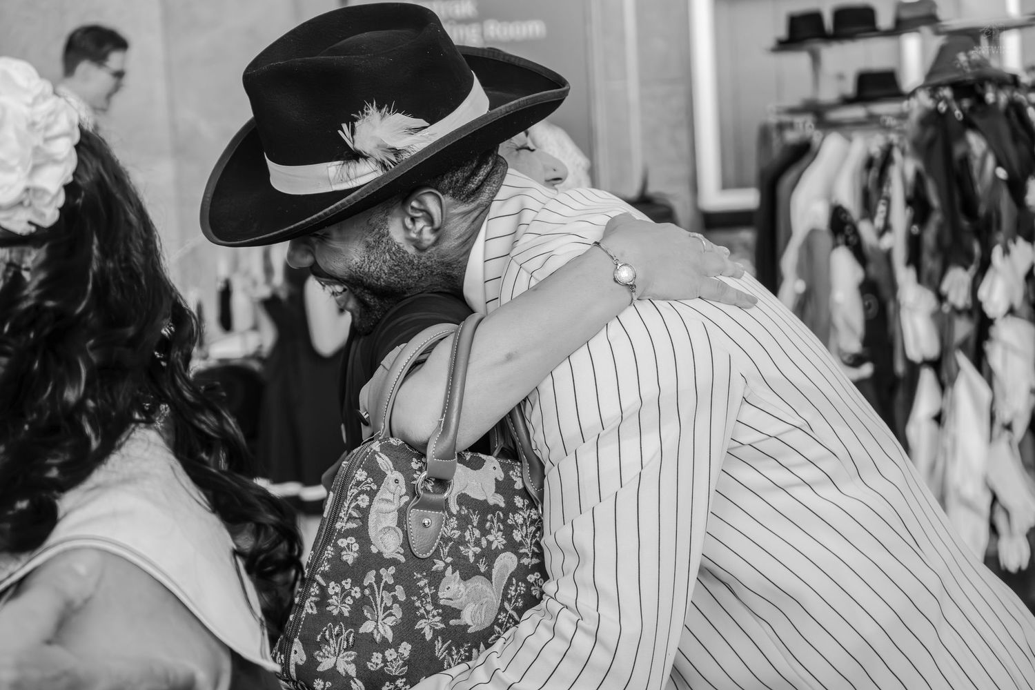 Black and white photo of two people embracing warmly in a clothing boutique, one wearing a wide-brim hat and pinstripe jacket.