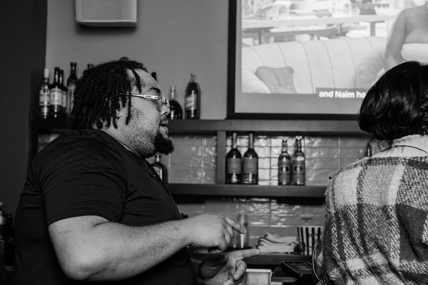 Black and white photo of two people conversing at a bar with bottles and a screen in the background.
