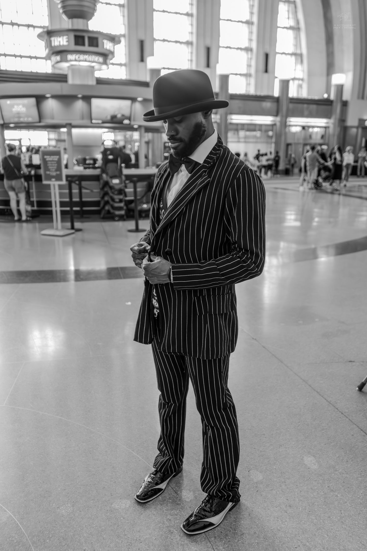 Stylish man in pinstripe suit and bowler hat poses in a busy airport terminal, black and white photo.