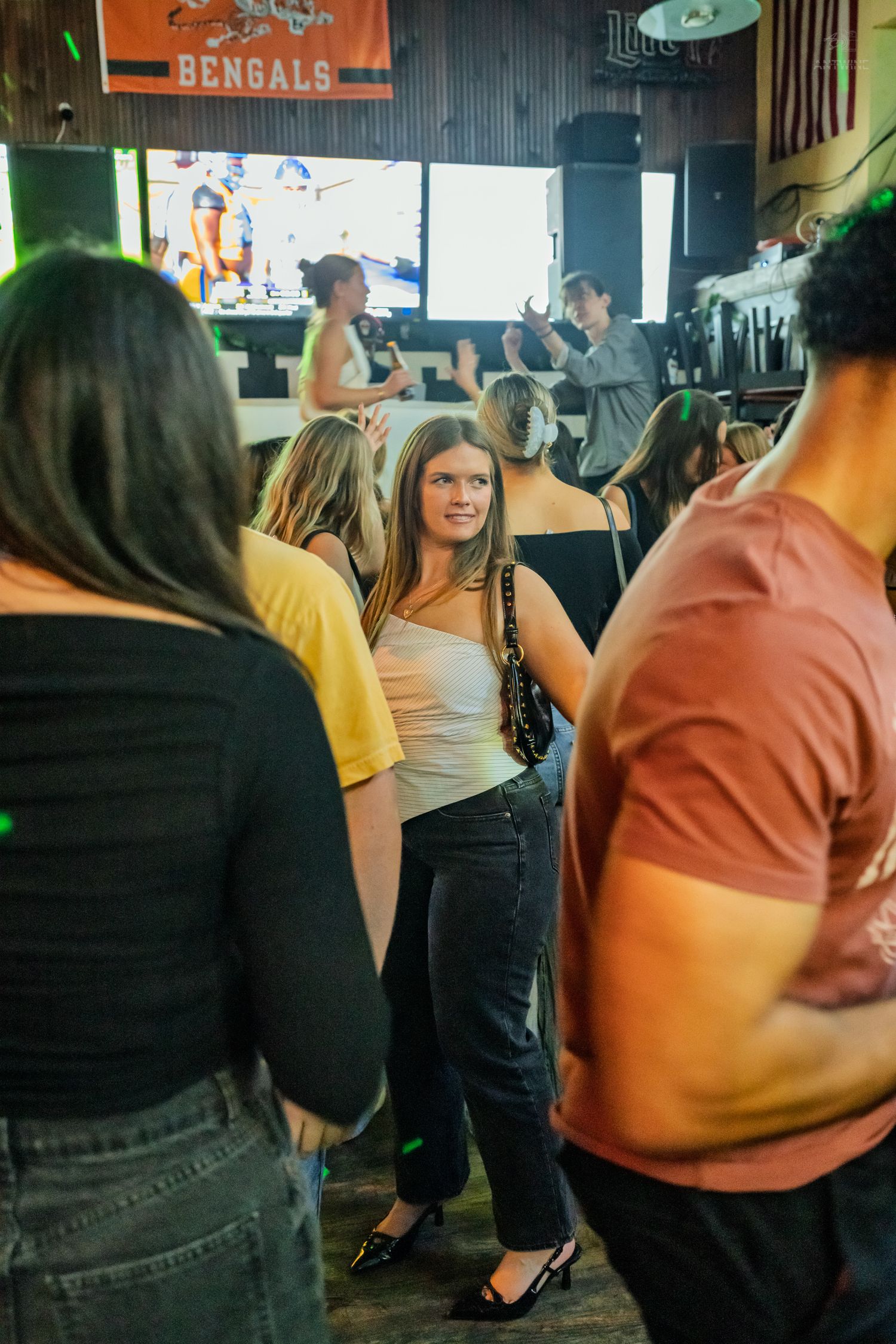 Crowded sports bar with Bengals banner, people socializing, woman in white top and black jeans smiling at camera.