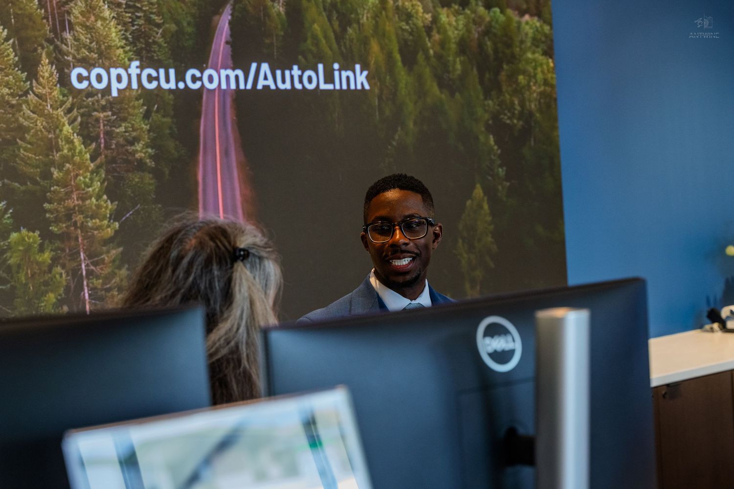 A smiling professional man in a suit sits at a desk in front of a copfcu.com/AutoLink advertisement screen.