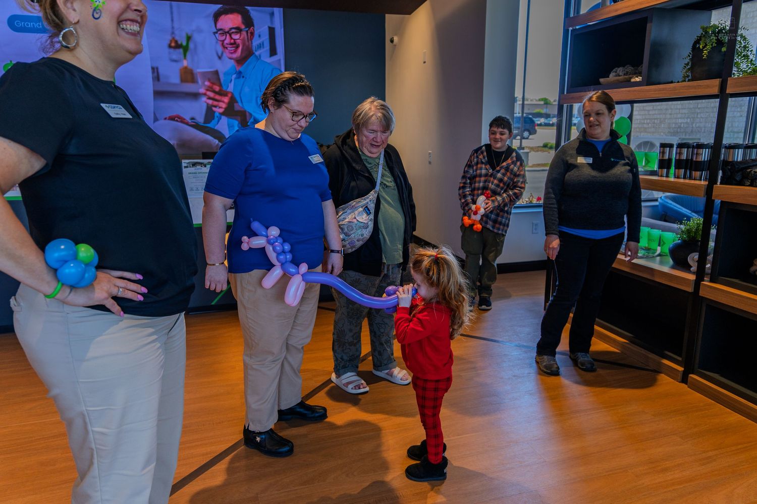 Credit union staff member presents a balloon animal to an excited toddler at a grand opening celebration.