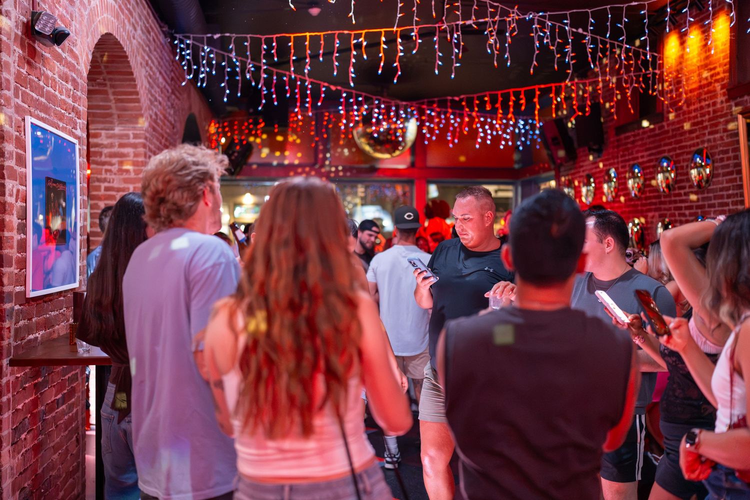 Crowd of people socializing inside a vibrant bar with colorful string lights and brick walls.