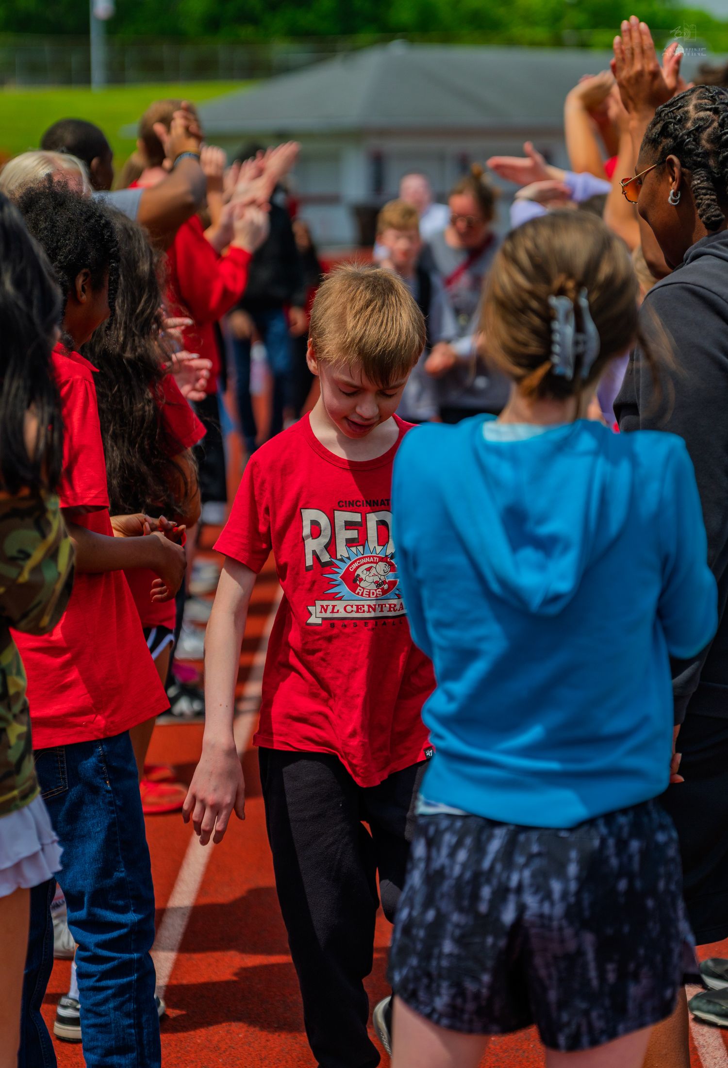 Smiling boy in red shirt walks through high-five tunnel of cheering peers on school track.