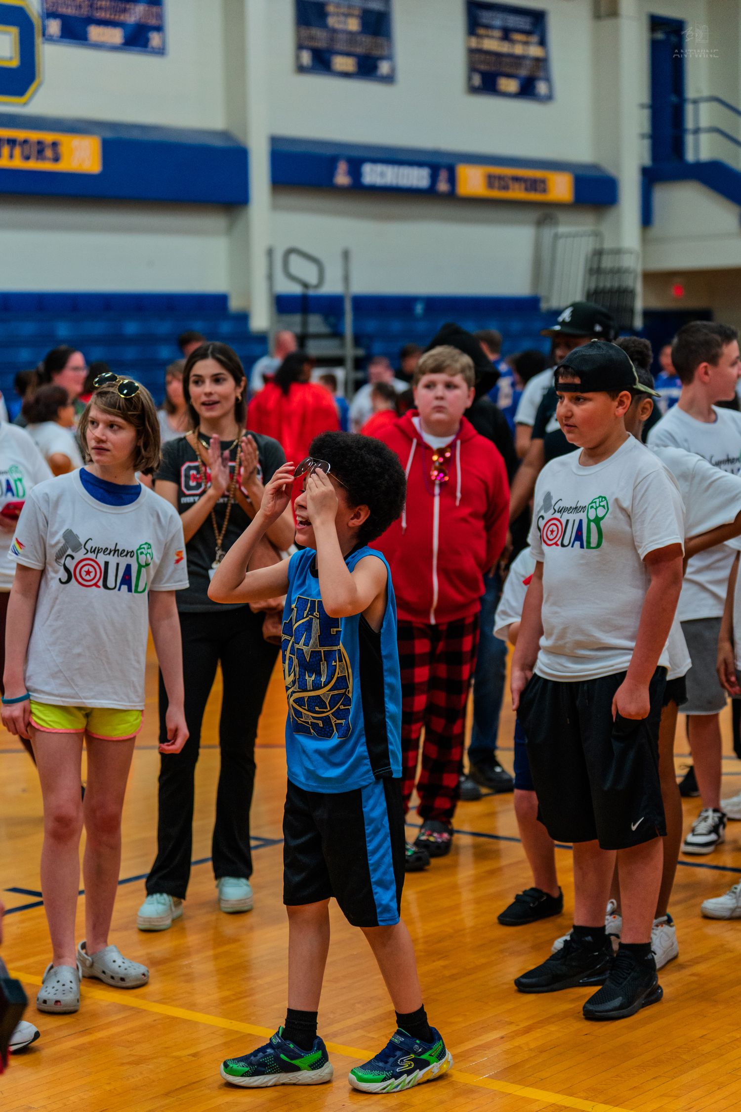 Children in Sunshine Squad shirts stand on a gym floor during a school event, with one boy shielding his eyes.