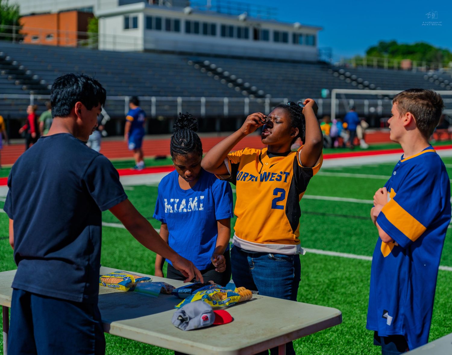 Four students stand around a table at a school field day event on a football field, one drinking from a cup.