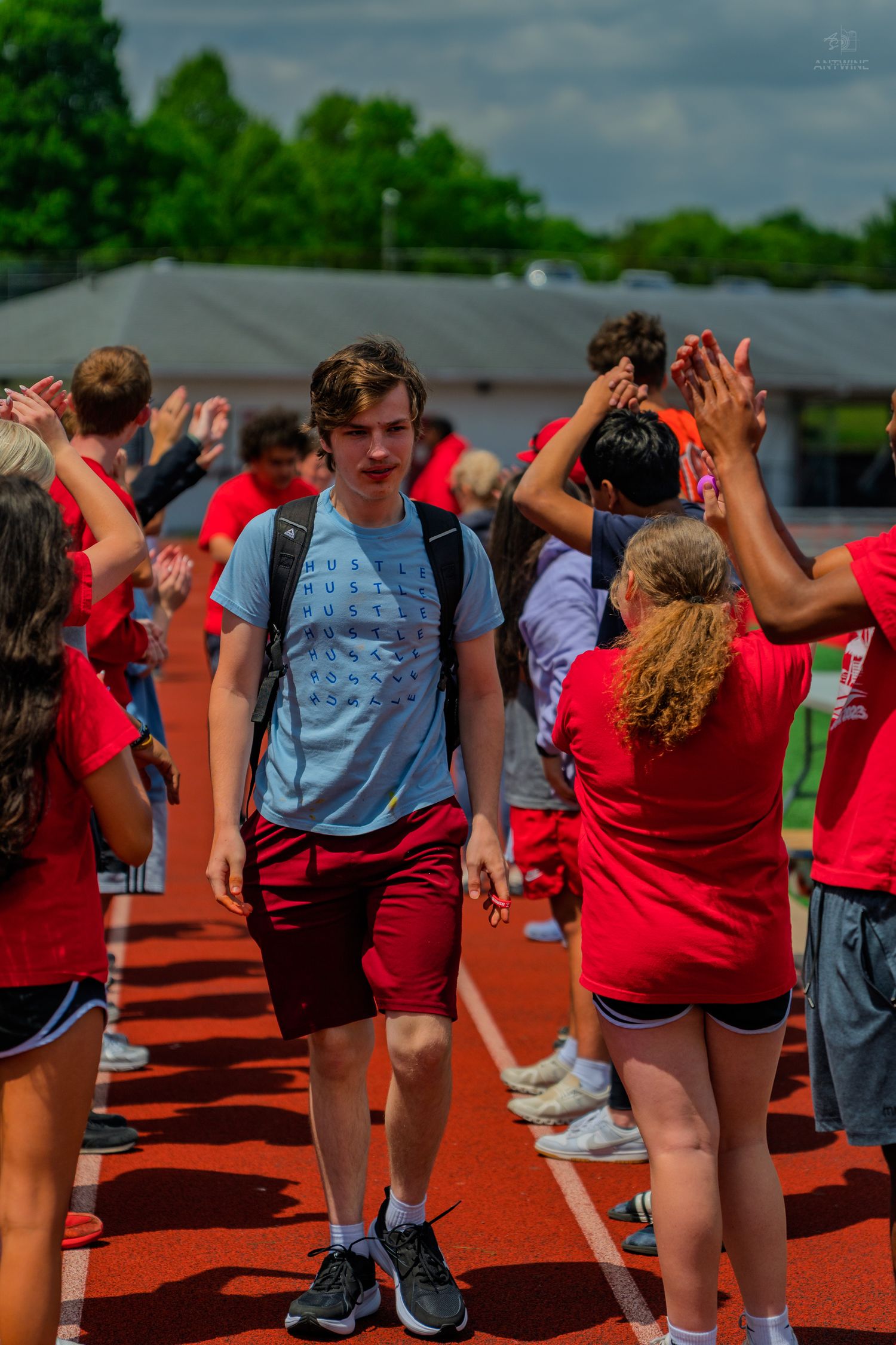 Students in red shirts high-five a teen with a backpack walking along a track at a school outdoor event.