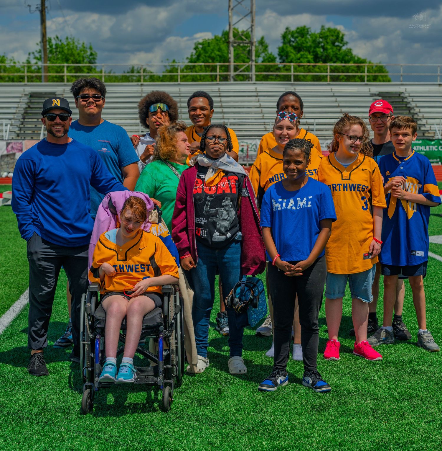 Group of diverse youth and adults posing together on a green sports field, including a student in a wheelchair.