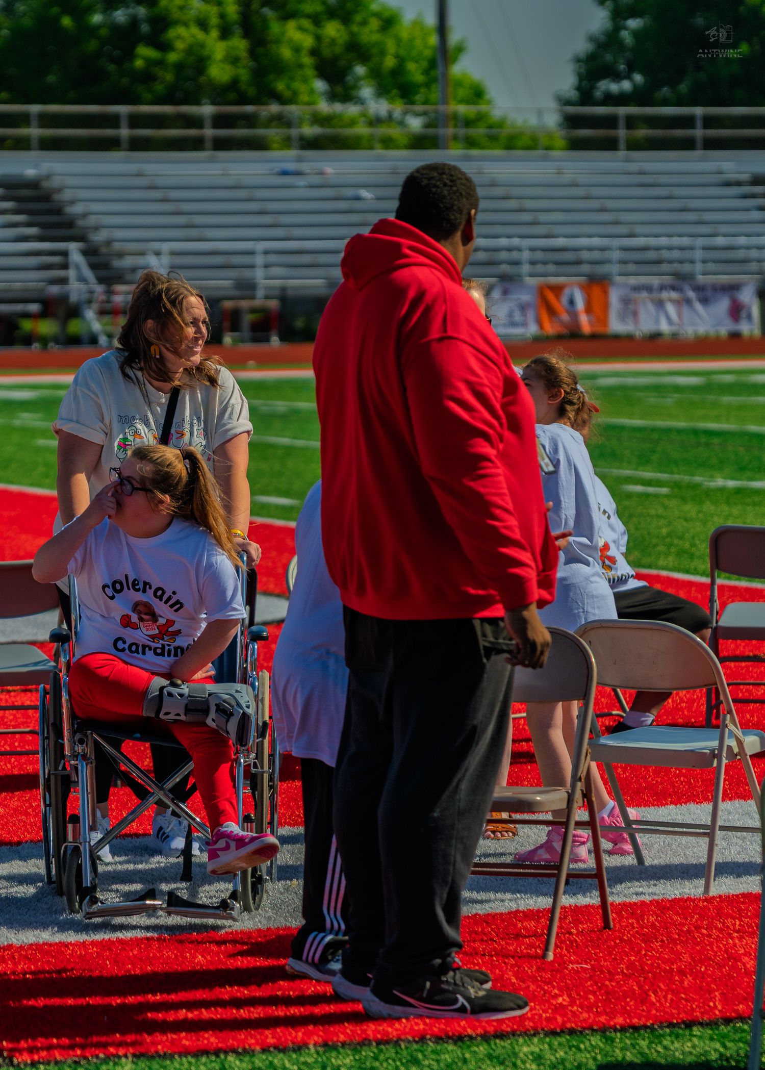 People in Colerain Cardinals shirts gather on a red football field, including a person in a wheelchair.