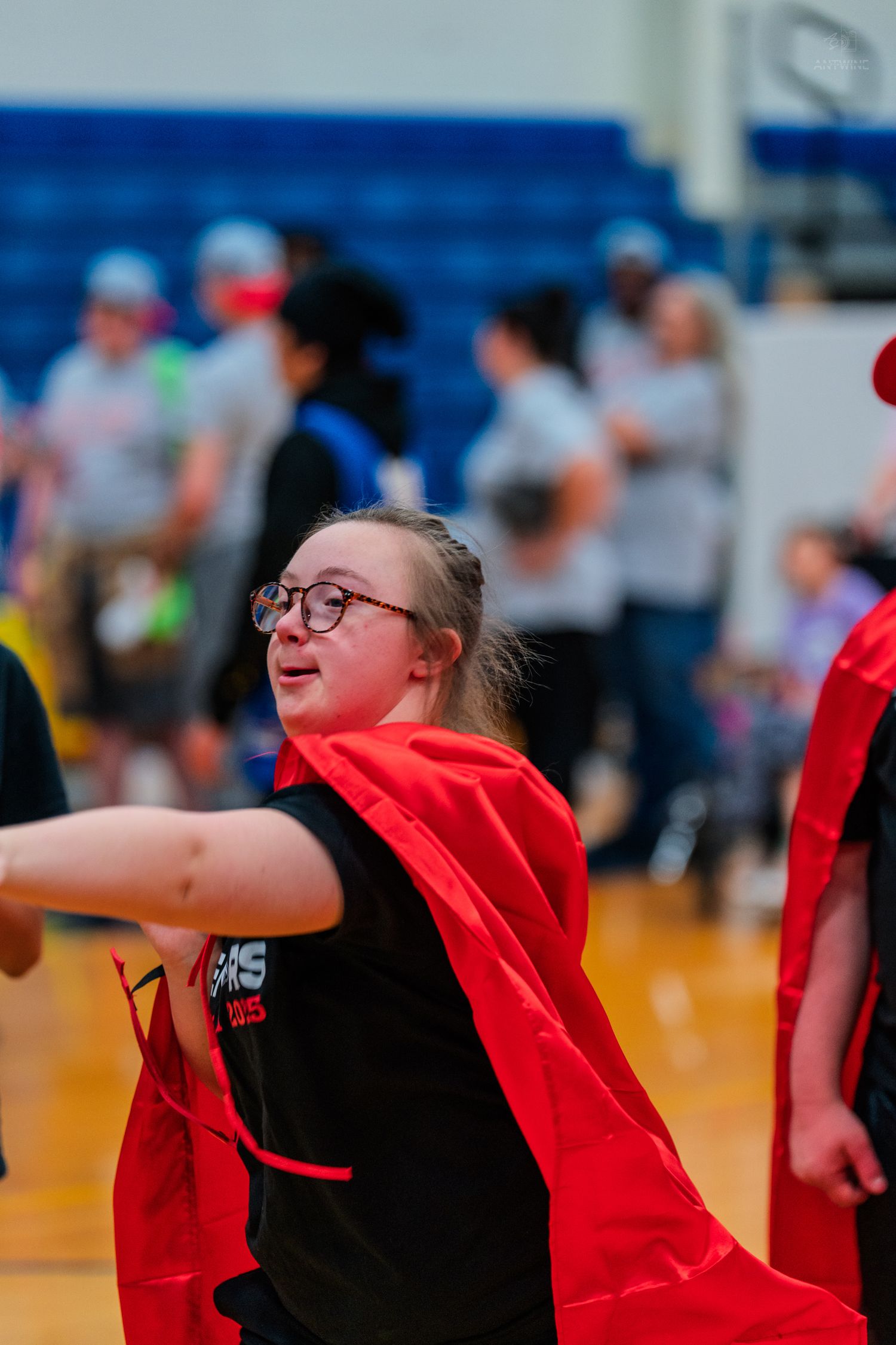 Young girl with glasses wearing a red cape participates in a Special Olympics basketball event in a school gymnasium.
