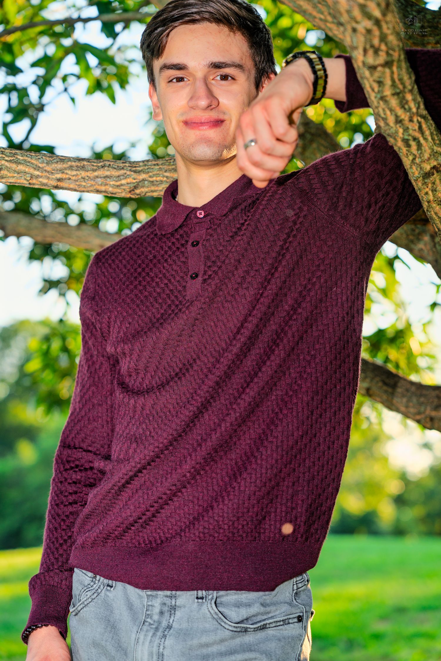 Young man in burgundy polo sweater leaning against tree in park, smiling outdoors.