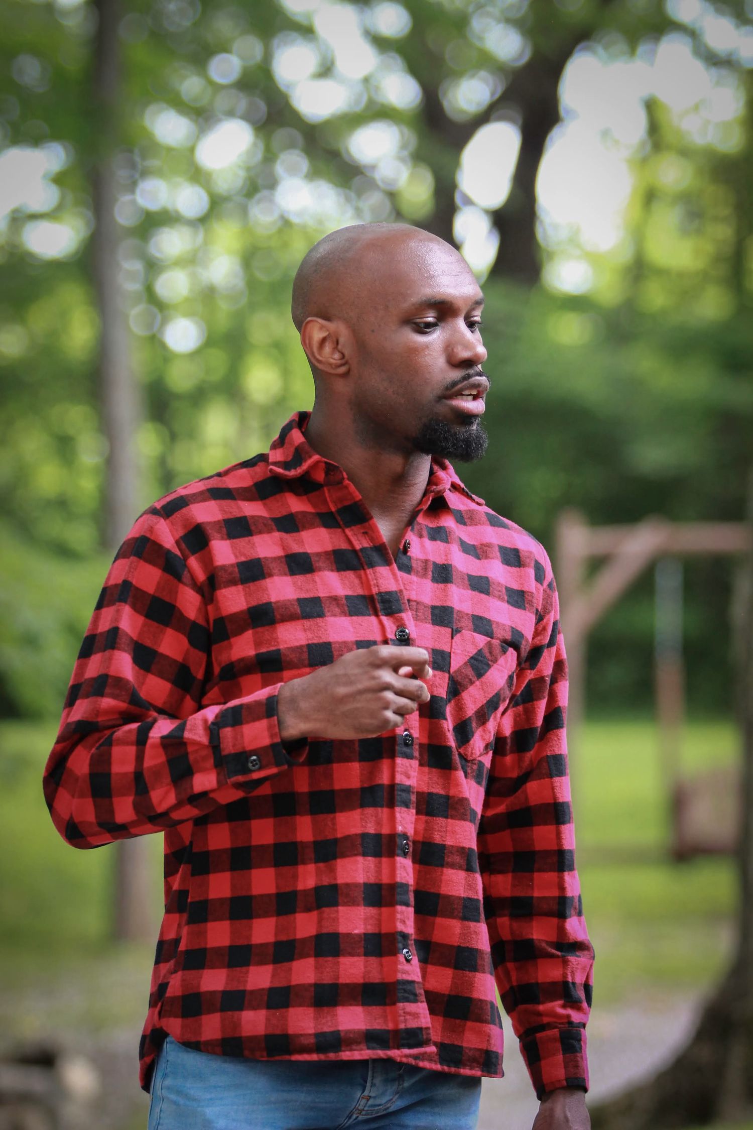 Bald Black man with beard wearing red plaid flannel shirt and jeans standing outdoors in green park setting.