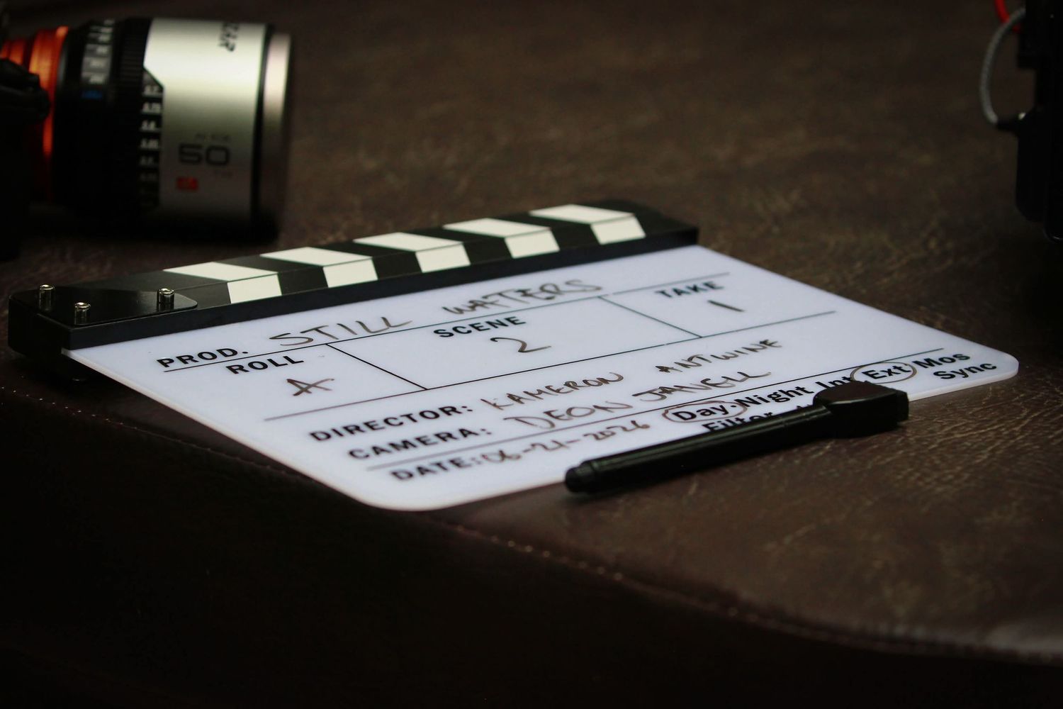 A film production clapperboard resting on a dark surface next to a camera lens on a movie set.