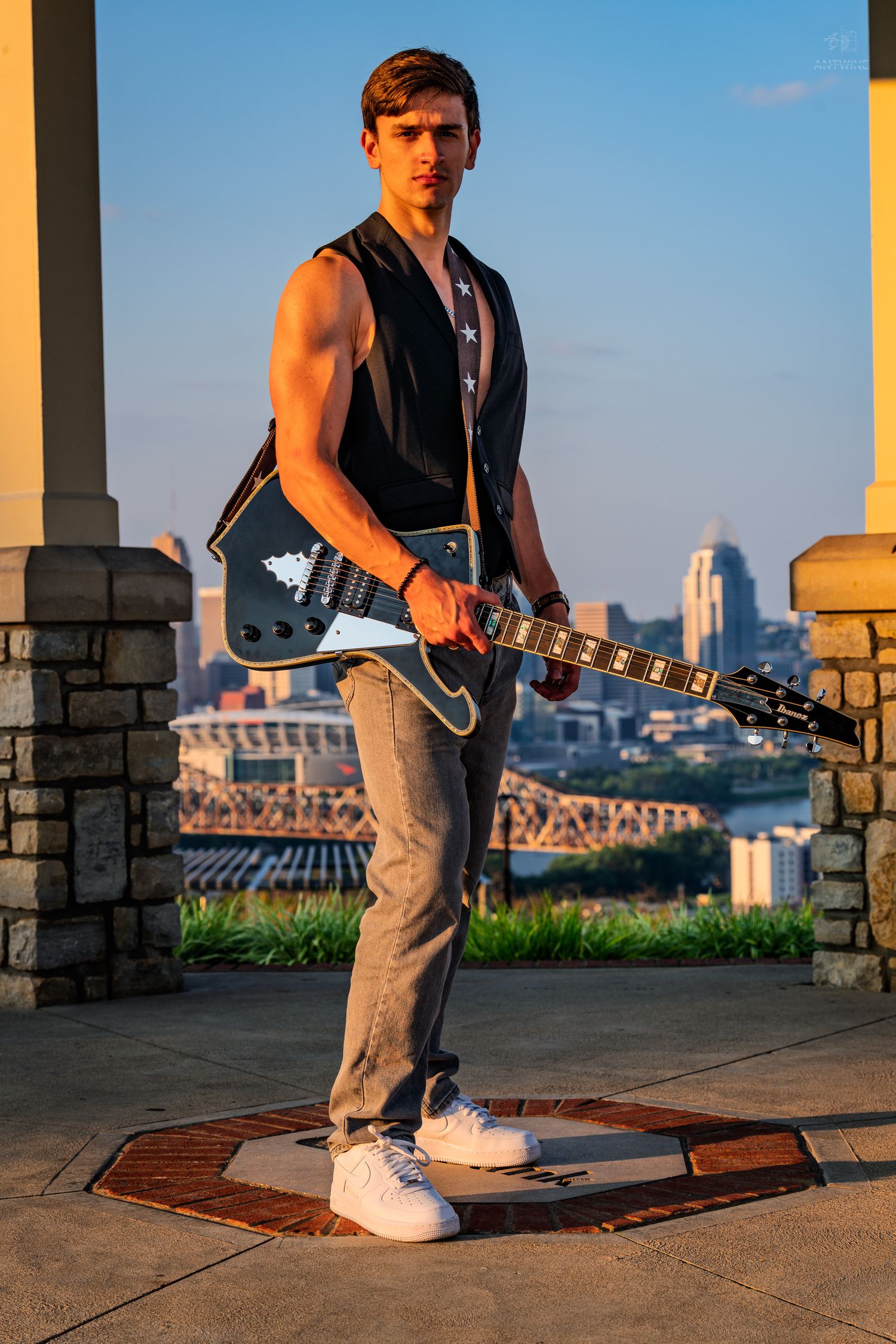Young muscular guitarist holding electric guitar outdoors at golden hour with city skyline backdrop.