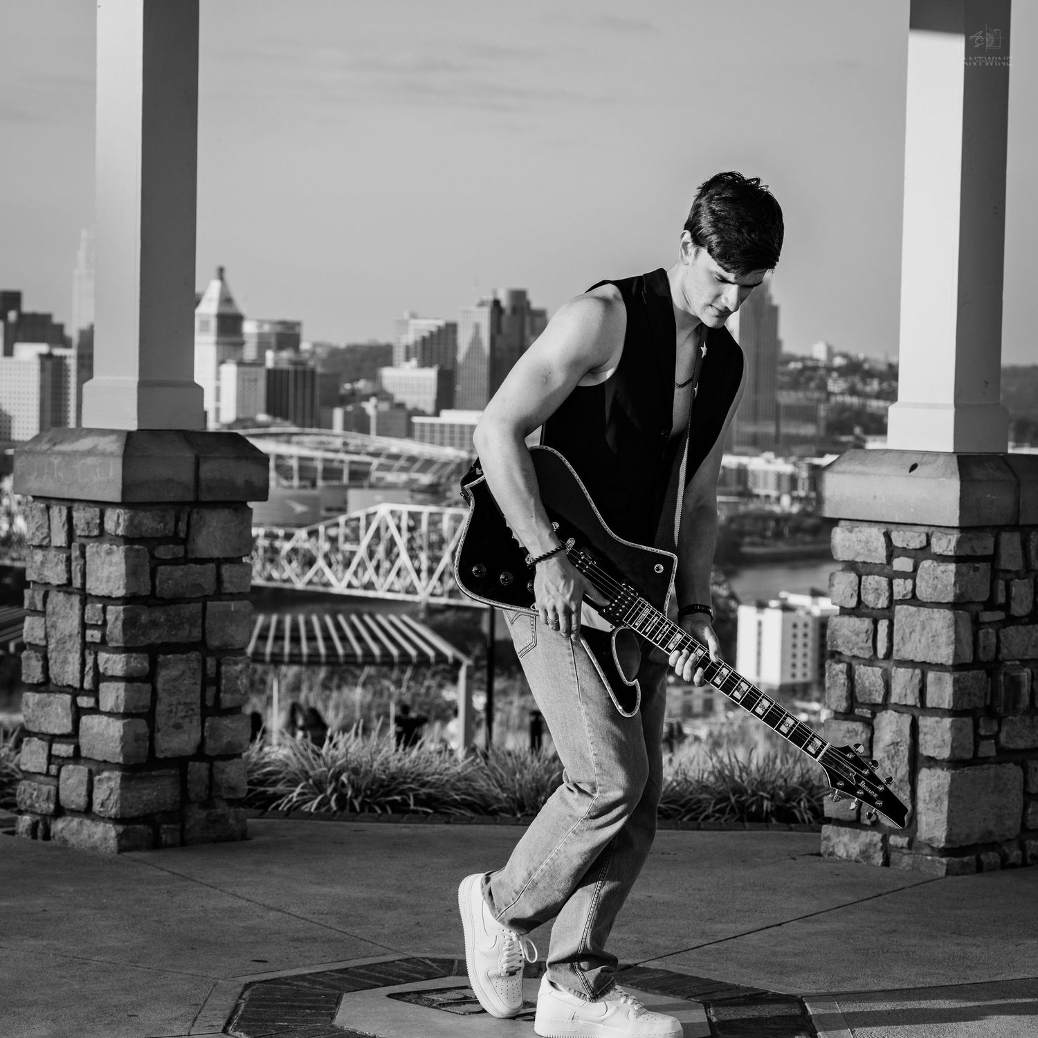 Black and white photo of a young man playing electric guitar outdoors with a city skyline in the background.