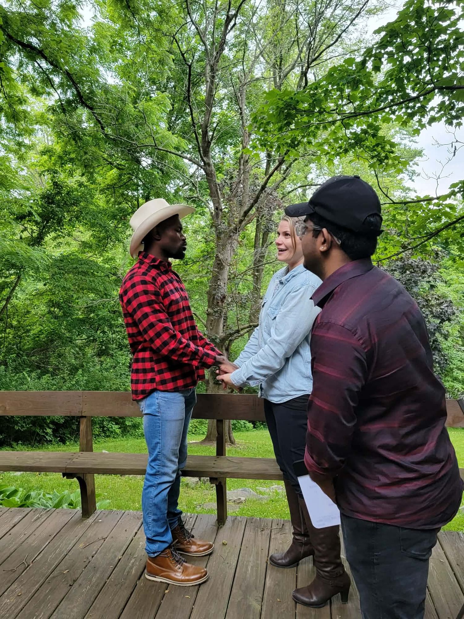 Three people conversing on wooden deck surrounded by lush green trees.