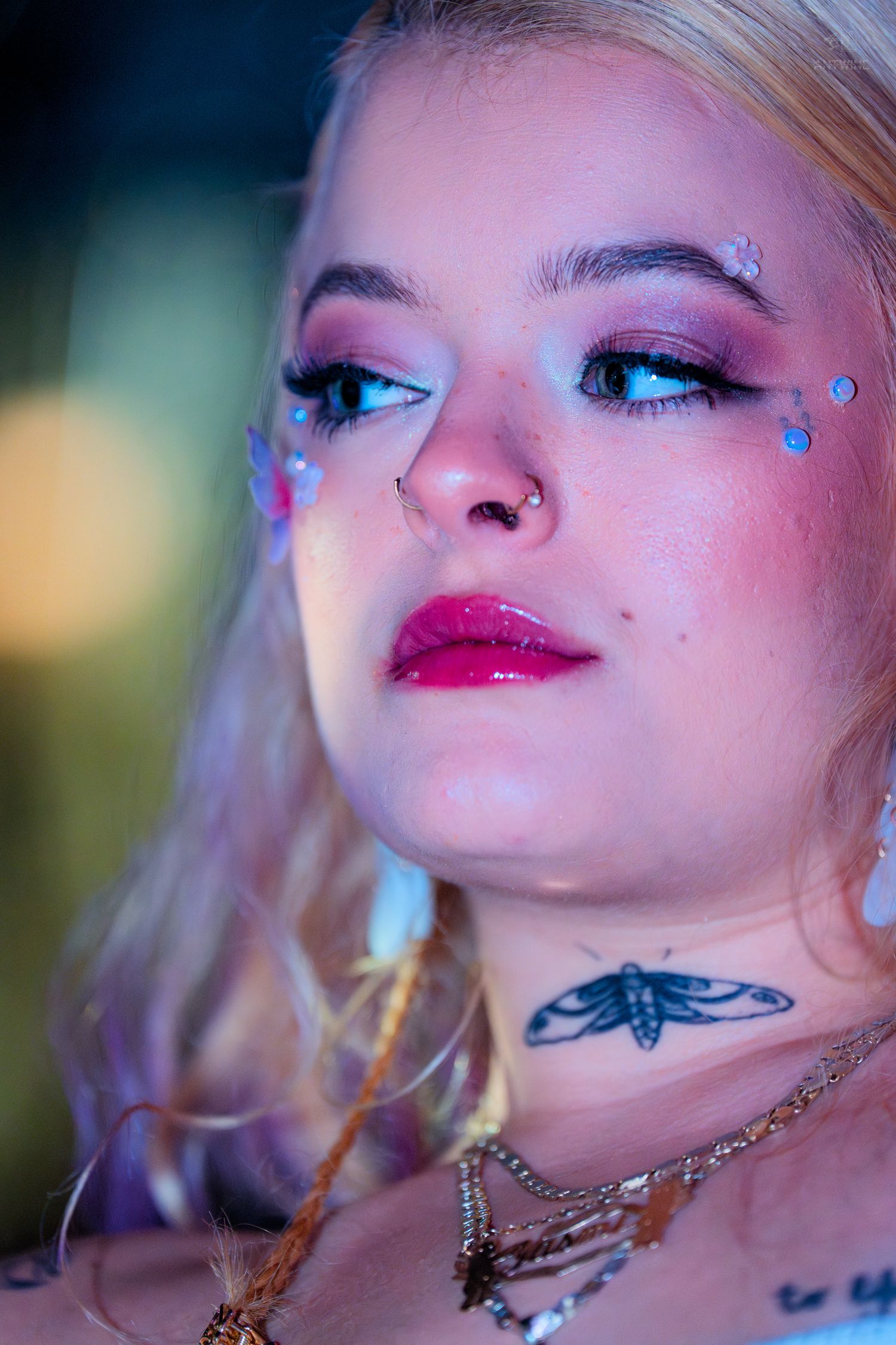 Portrait of blonde woman with colorful eye makeup, glitter gems, red lips, and moth neck tattoo under neon light.