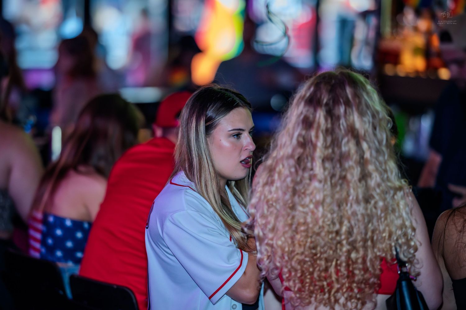 Two women having a conversation at a busy bar with colorful background lights.
