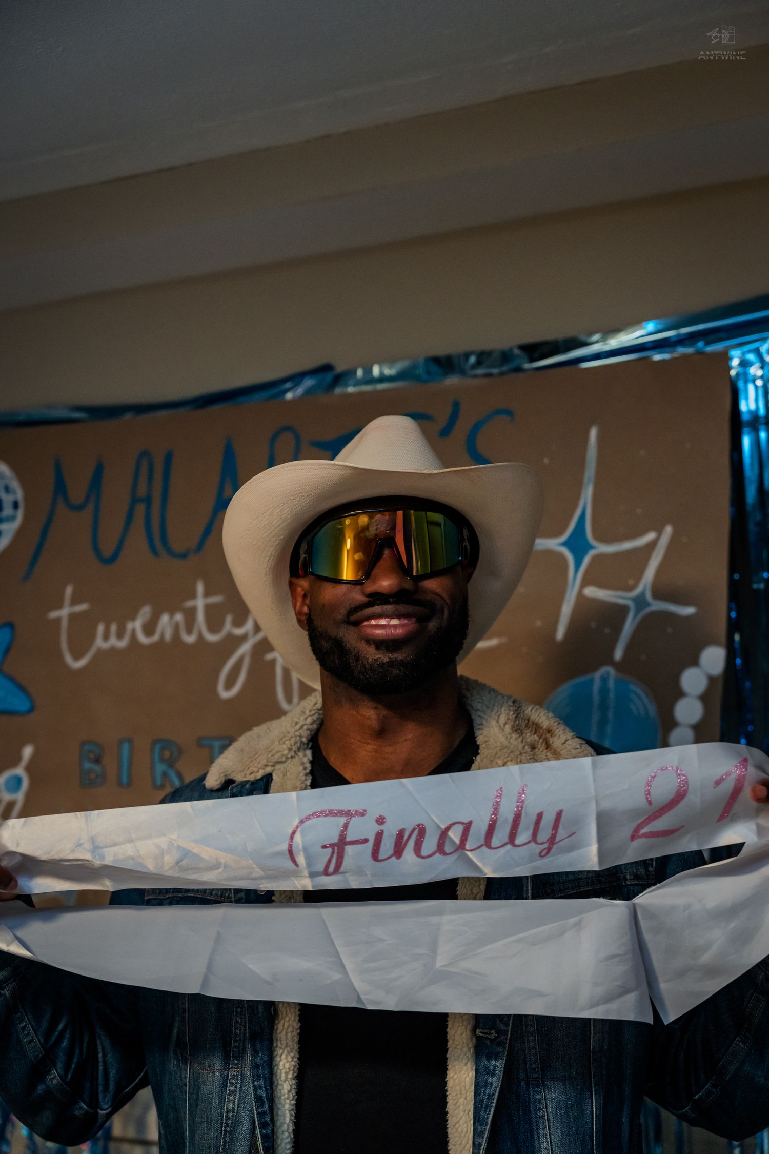 Man smiling in white cowboy hat and mirrored sunglasses holding a 'Finally 21' birthday sash.