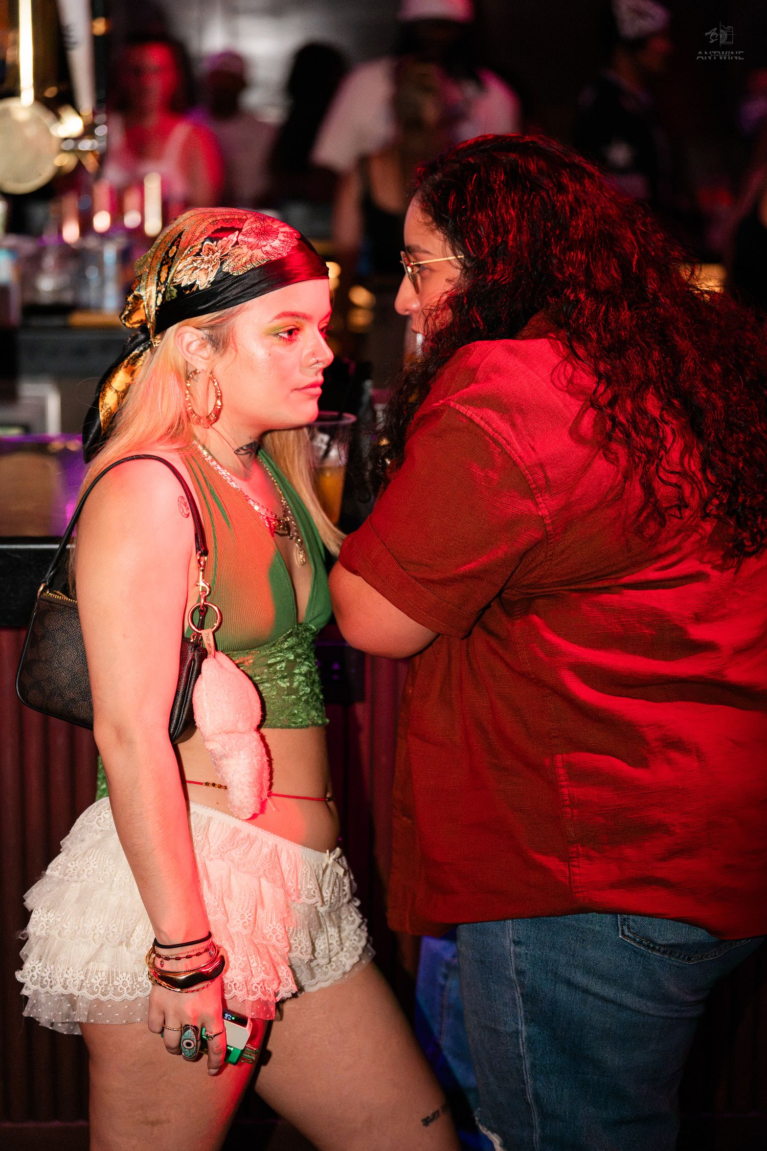 Two women in festive costumes share an intimate conversation at a vibrant nightclub bar.