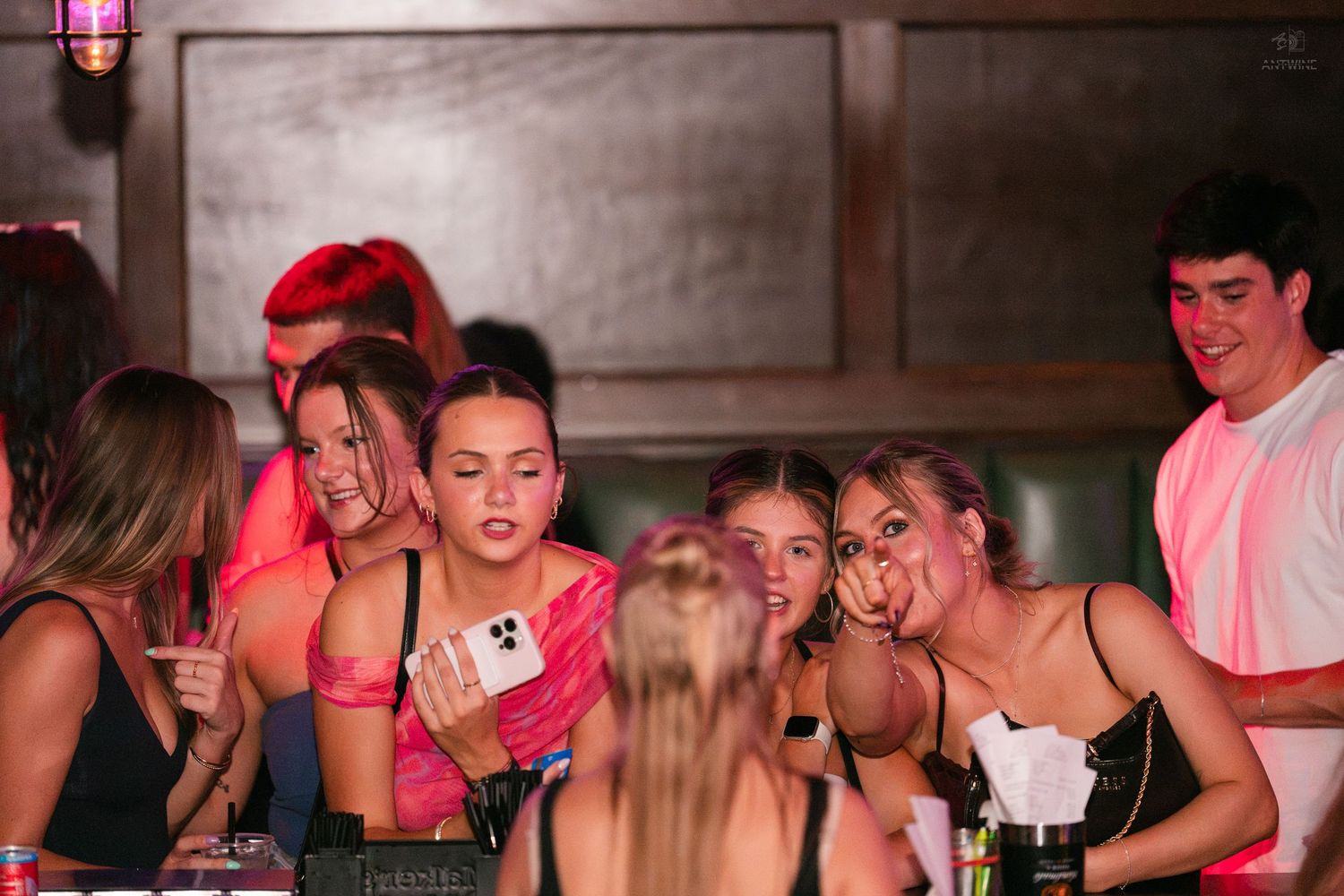 Group of young women huddle together looking at a phone at a lively bar or nightclub.
