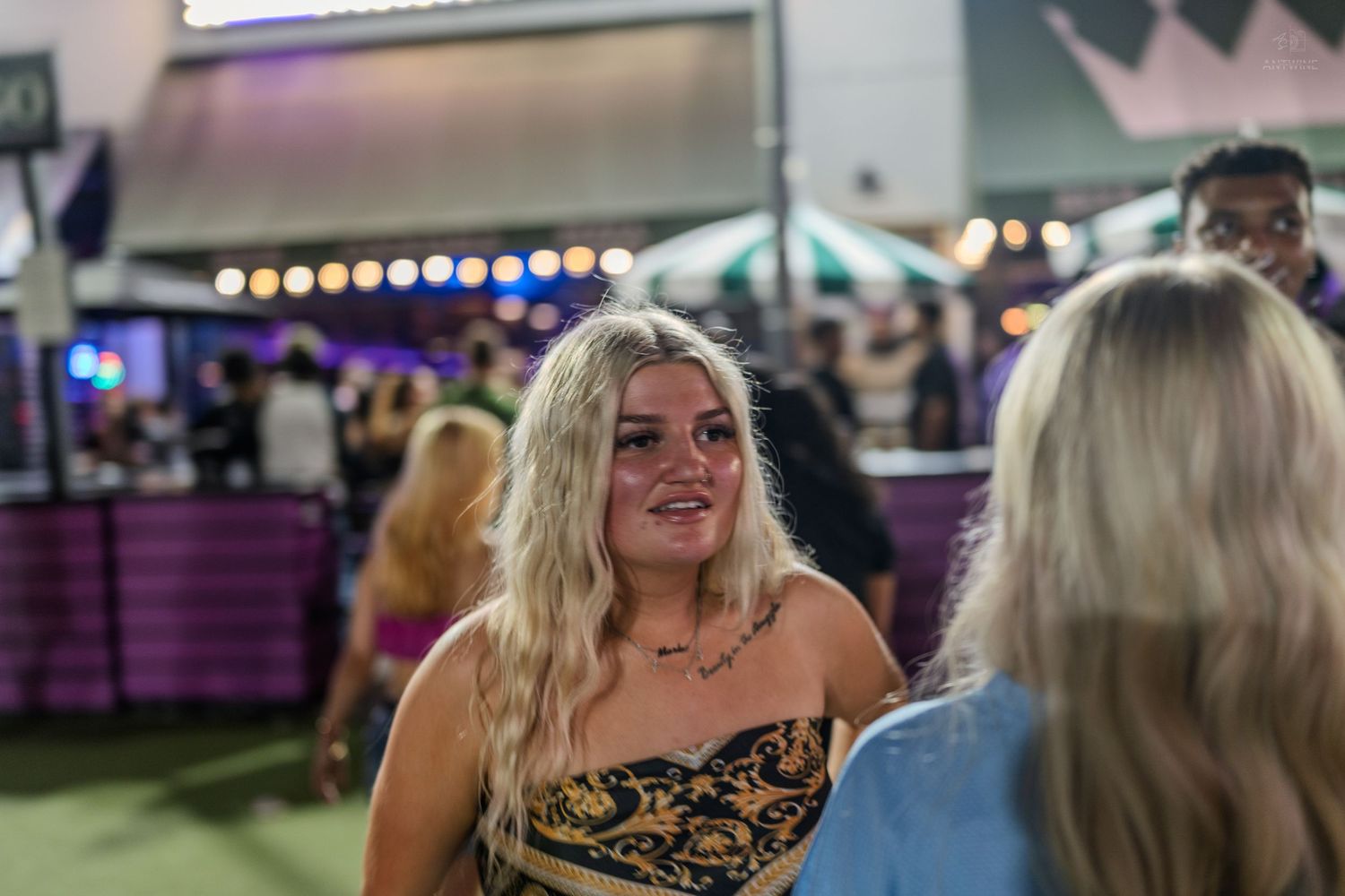 Young blonde woman in floral top smiling at an outdoor evening event with purple lights.