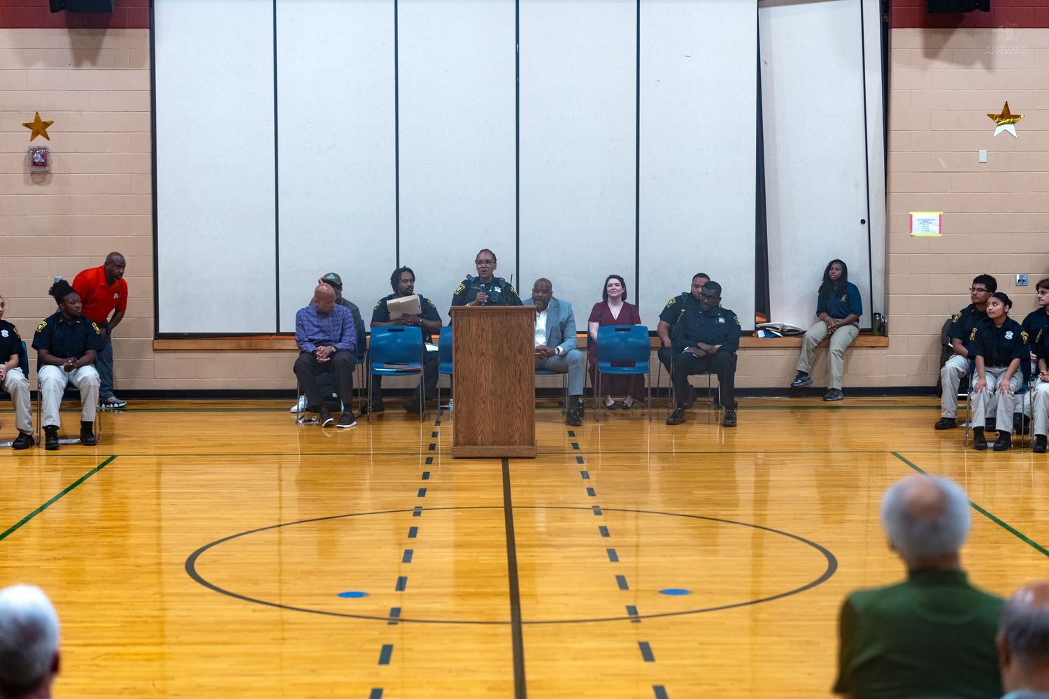 Community meeting in a school gymnasium with speaker at podium and attendees seated in chairs on a basketball court.