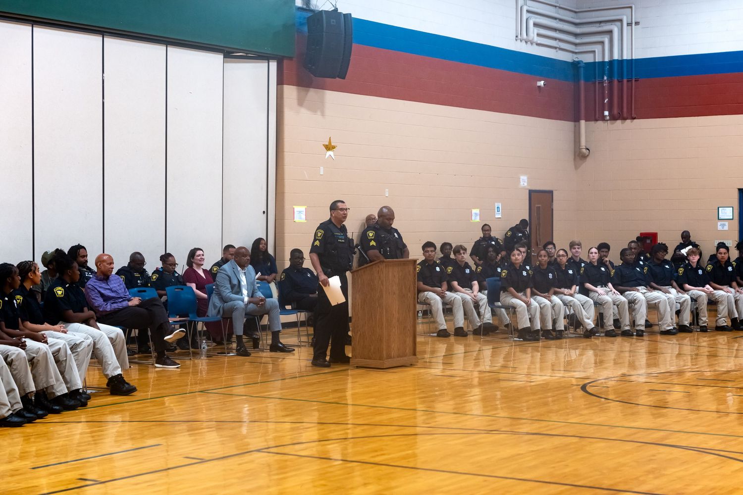 Police officers and students gathered in a school gymnasium for a formal assembly ceremony at a podium.