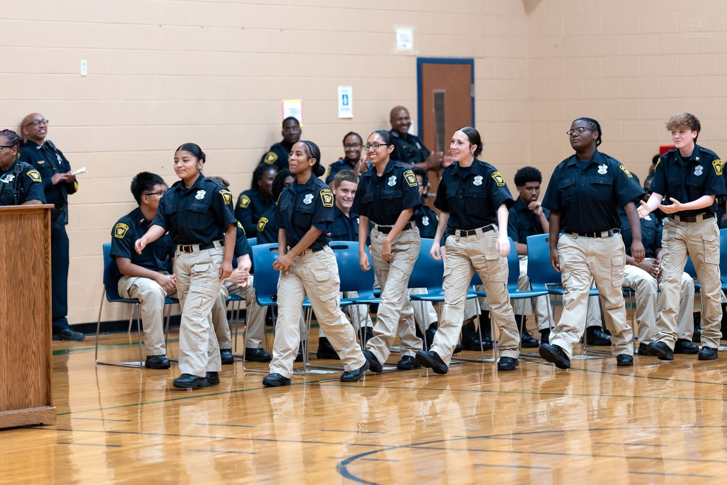 Young students in matching navy polo shirts and khaki pants stand in a school gymnasium during a formal ceremony or presentation.