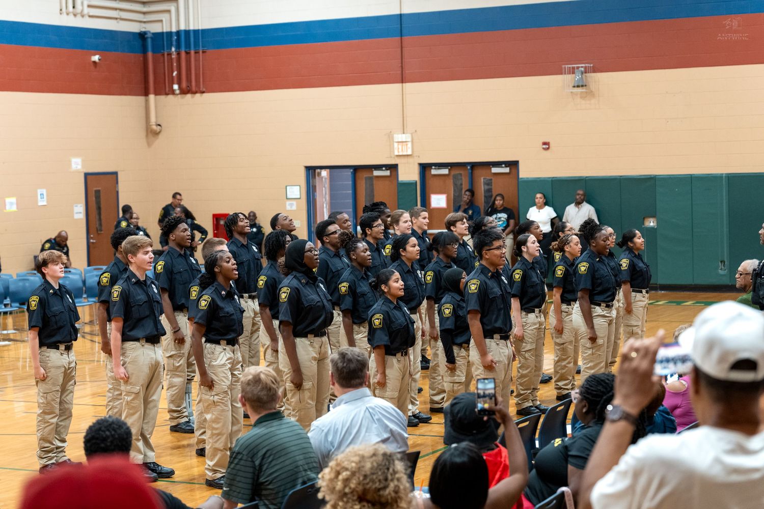 Police cadets in uniform stand in formation during an indoor ceremony in a school gymnasium with seated audience.